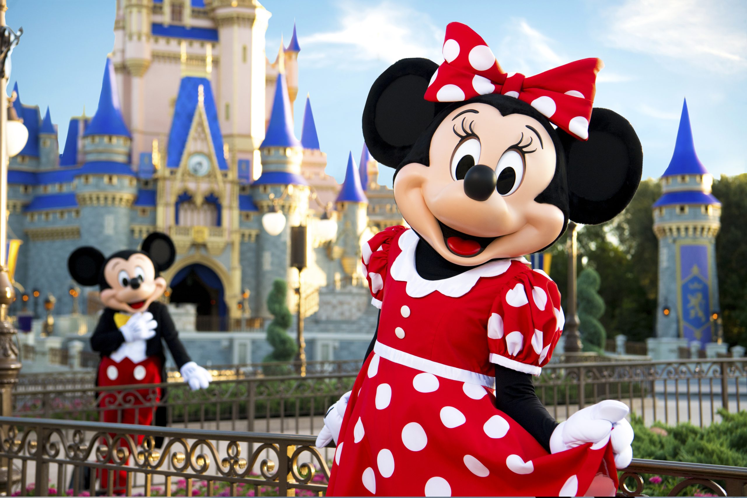 Minnie Mouse Character performer smiling while standing in front of Cinderella's Castle at the Magic Kingdom. Mickey Mouse looks on adoringly from the background