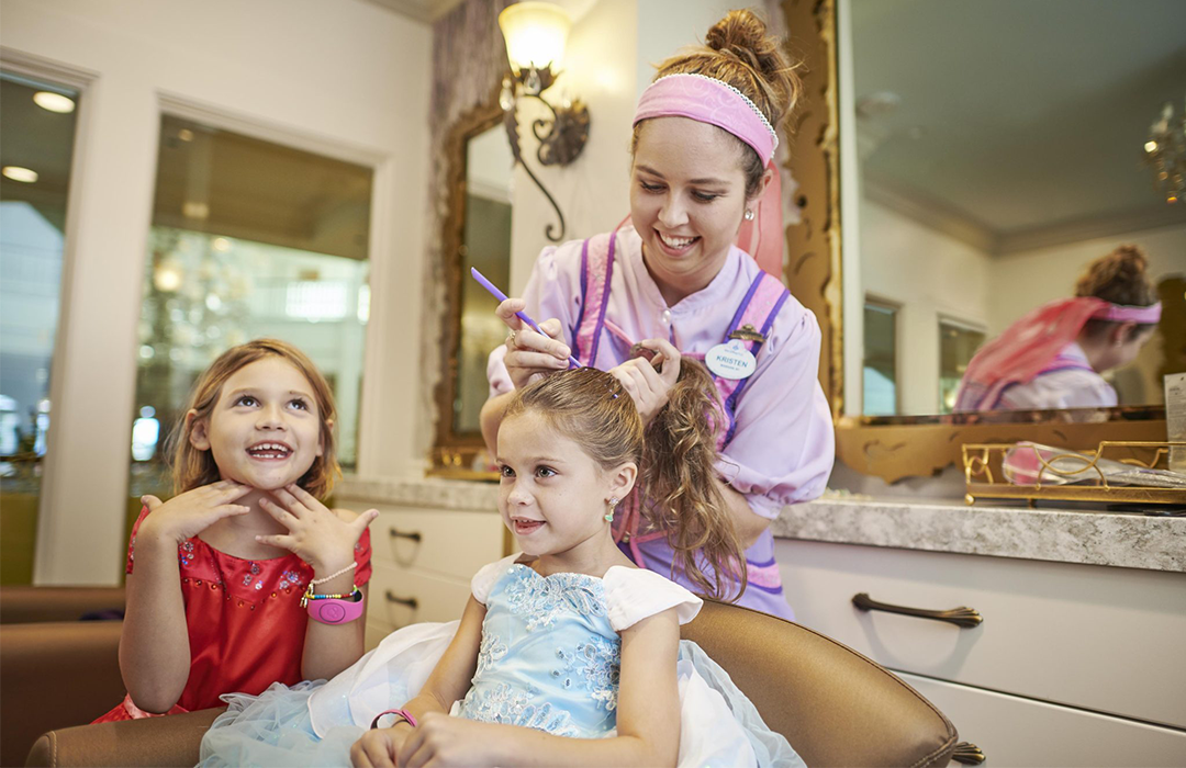 Two kids sitting in salon chairs smiling while a disney college program cast member styles their hair at the Walt Disney World Bibbidy Bobbidy Boutique