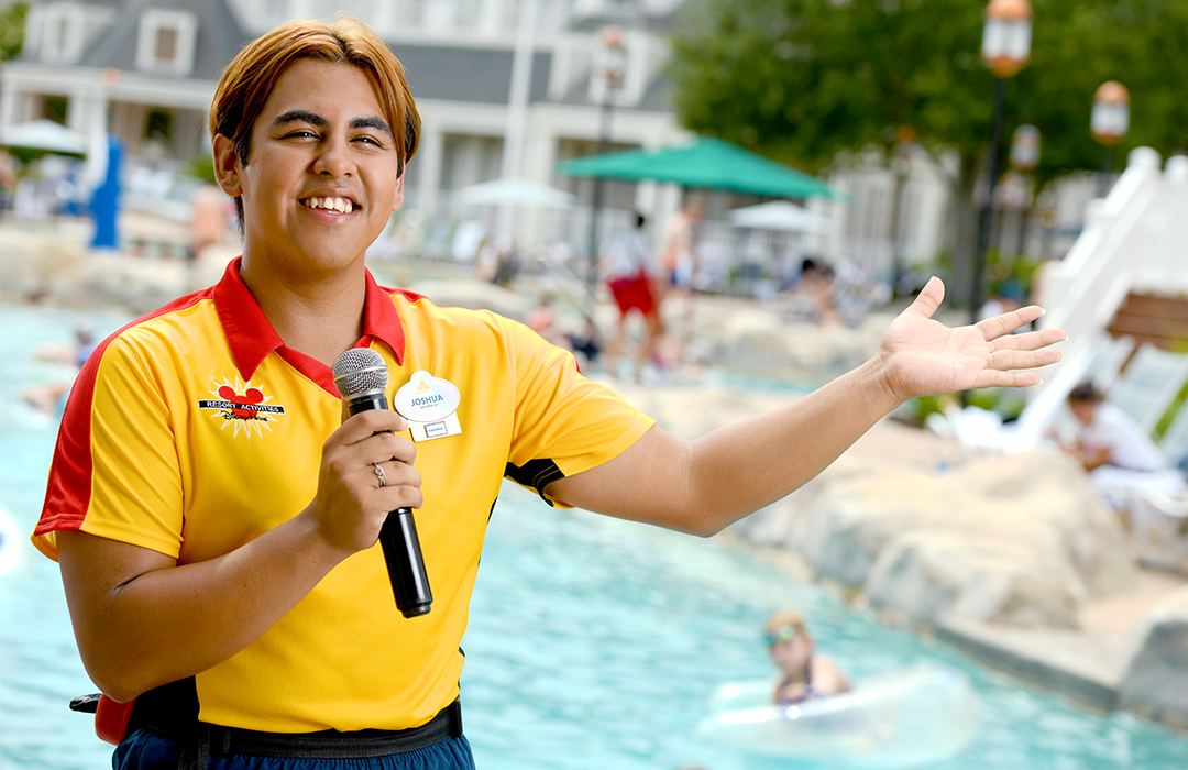 Disney college program cast member holding a microphone and smiling in front of a pool at a Walt Disney World Resort