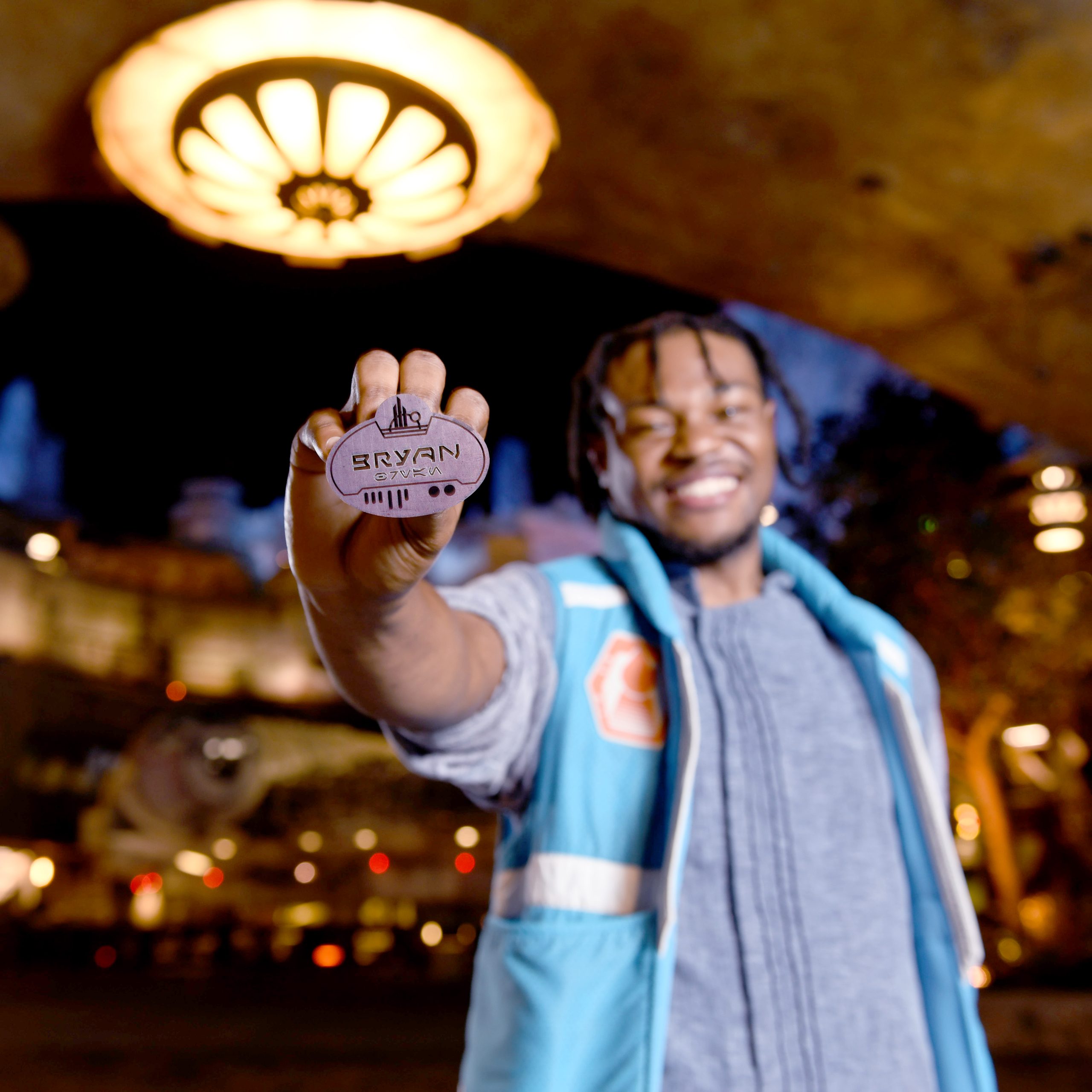 Male Disney College Program attractions participant smiling while holding his name badge forward towards the camera with Star Wars: Galaxy's Edge in the background