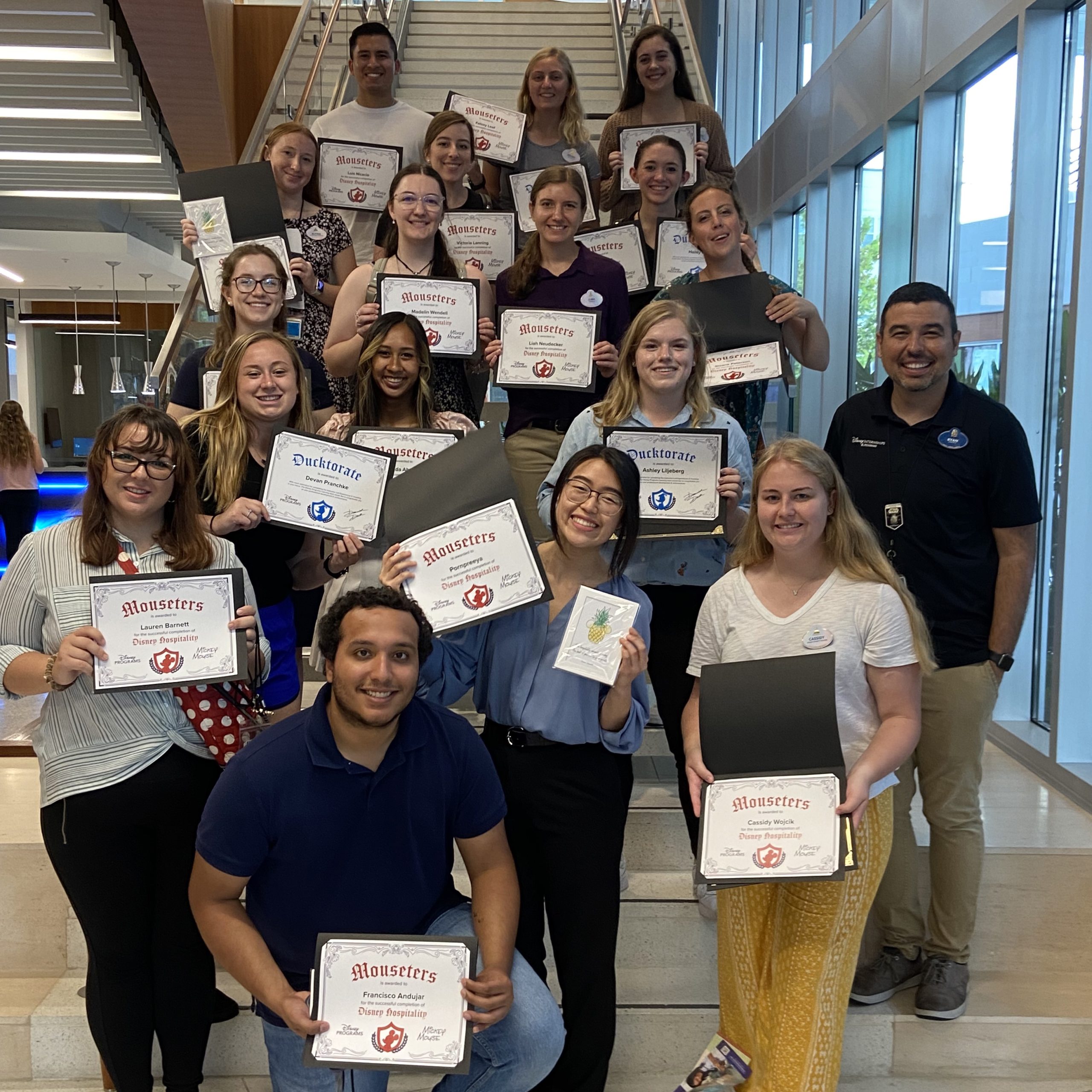 Eighteen Disney College Program participants standing on a stairwell smiling while holding up various certificates