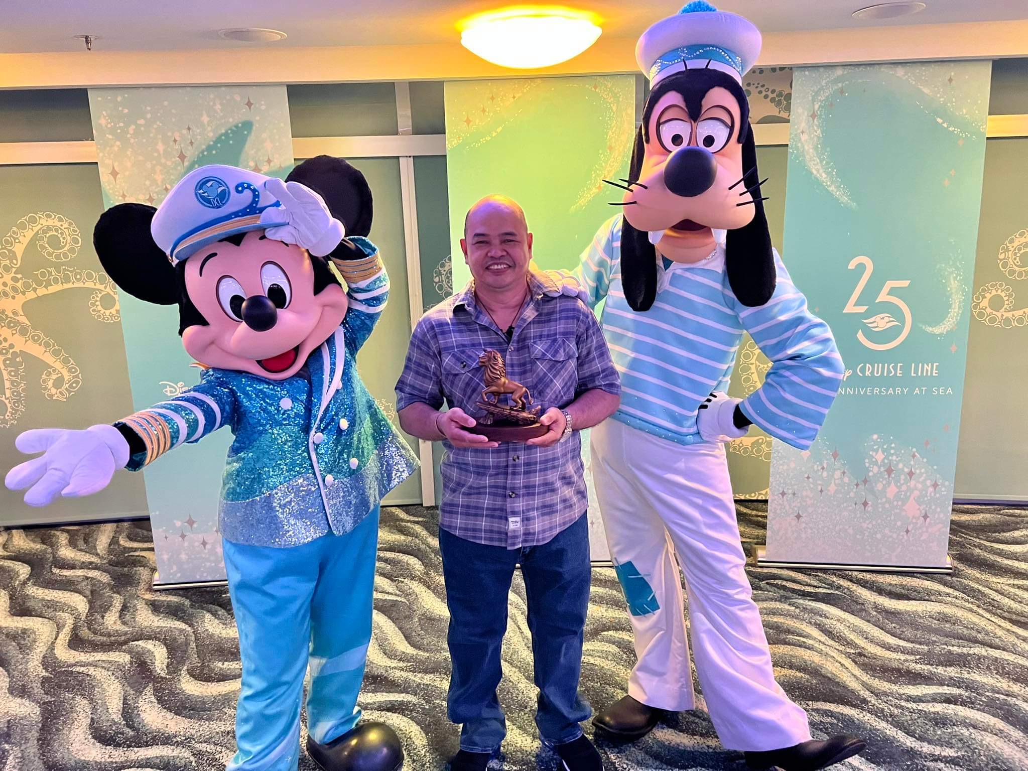 Ramil stands in between Mickey and Goofy in their 25-year anniversary costumes as he is presented with his 20-year company anniversary statue (statue has Simba from The Lion King posing on Pride Rock)
