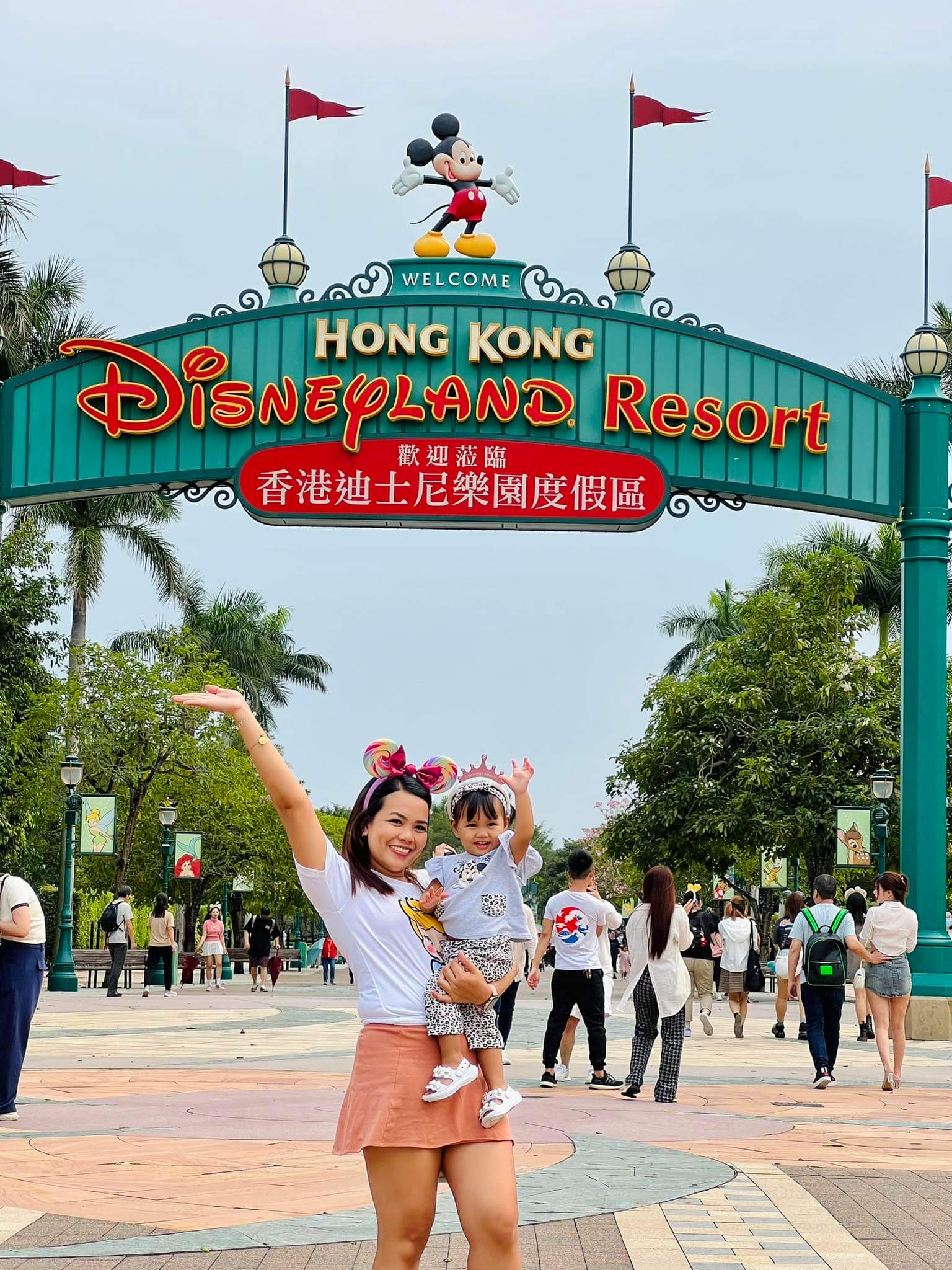 Leovy holds her daughter as they both raise their hands in excitement in front of the Hong Kong Disneyland Resort main entrance sign