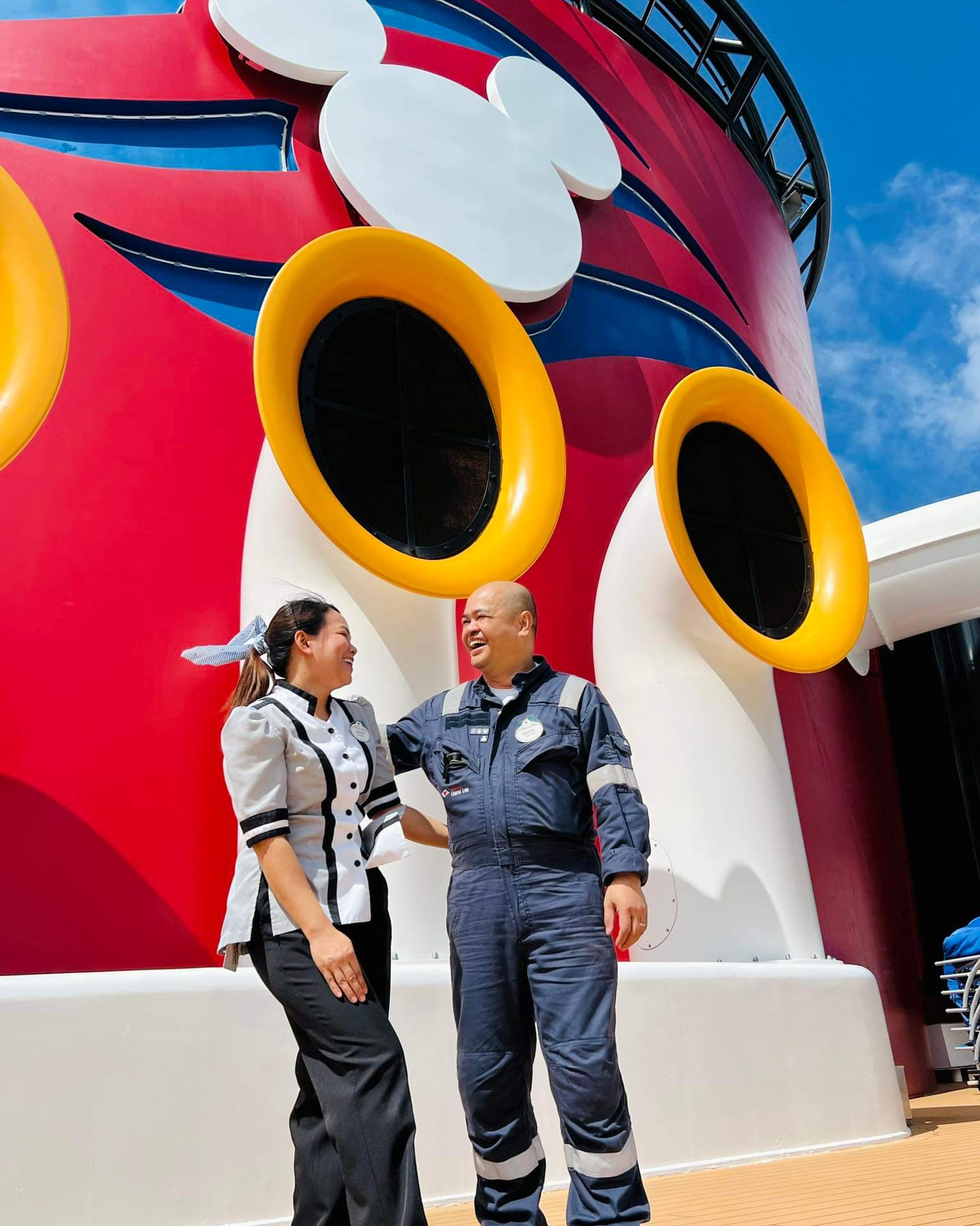 Leovy and Ramil, father and daughter, look at each other and laugh in their costumes on the Disney Fantasy in front of the iconic red, yellow and white funnel