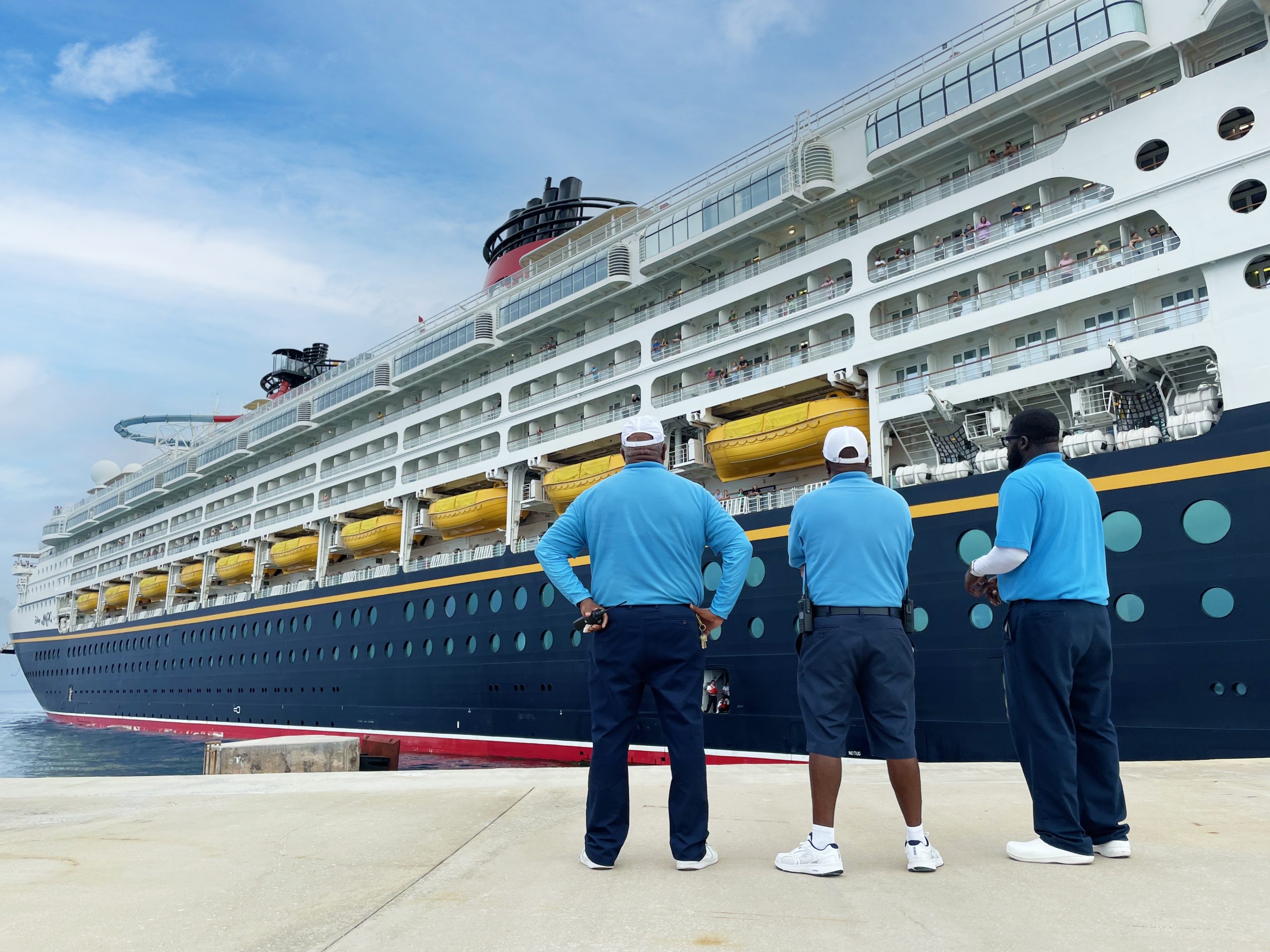 Three Disney Castaway Cay leaders stand with their backs to the camera looking at a Disney Cruise Line ship as it comes into port.