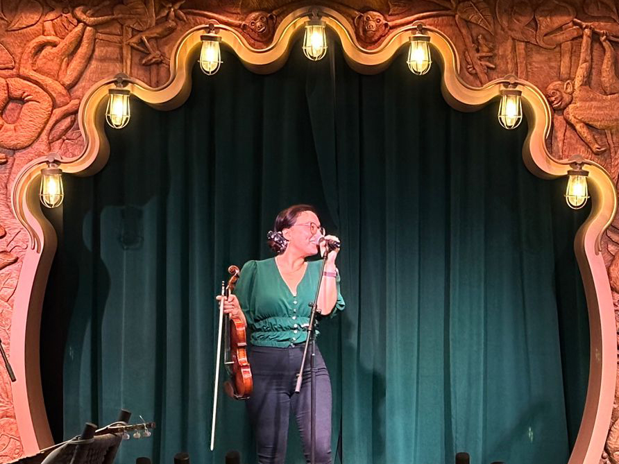 Erendira A mariachi musician in a green blouse performs with a violin and microphone on a decorated stage aboard the Disney Treasure.