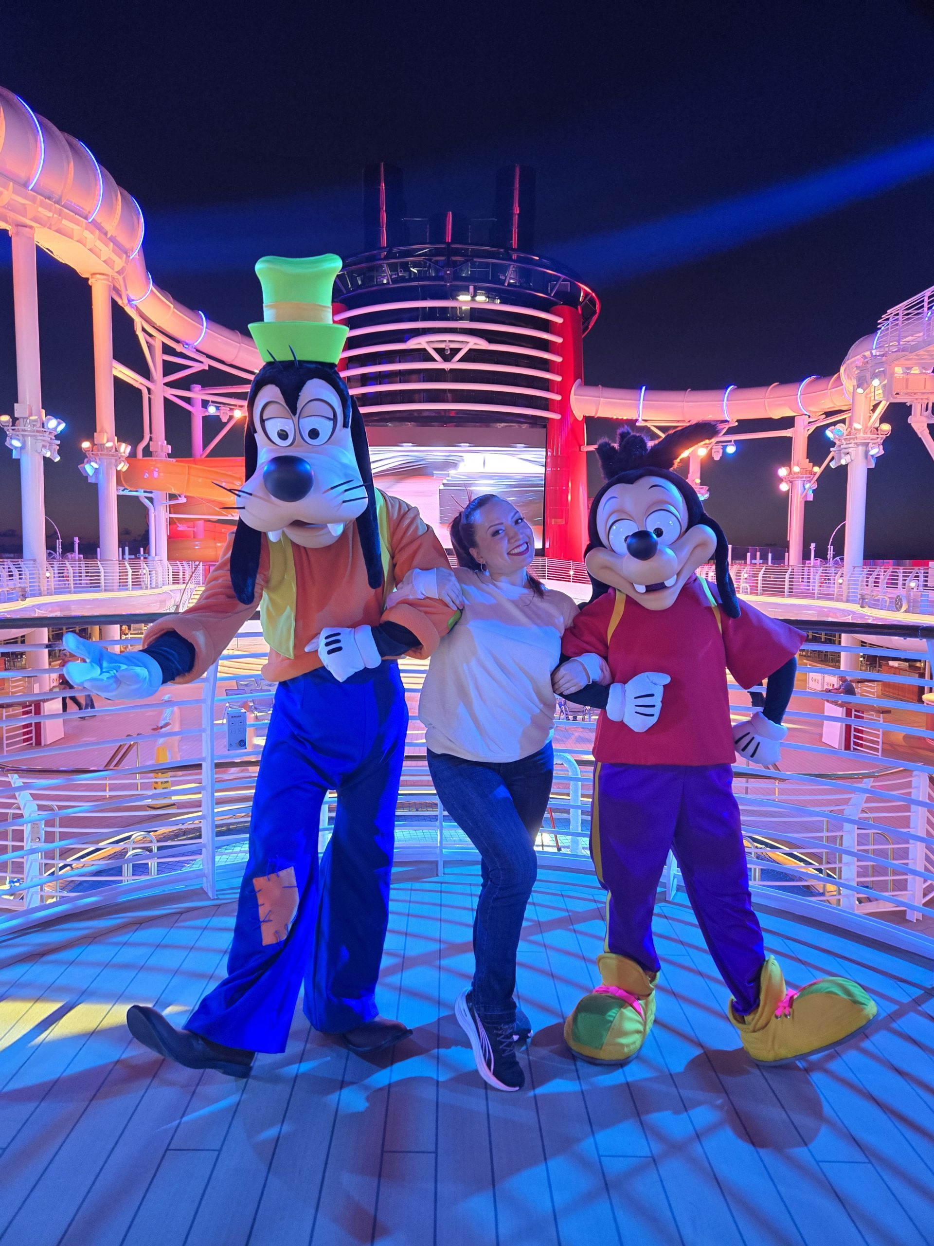A woman poses with Goofy and Max on a brightly lit cruise ship deck at night.