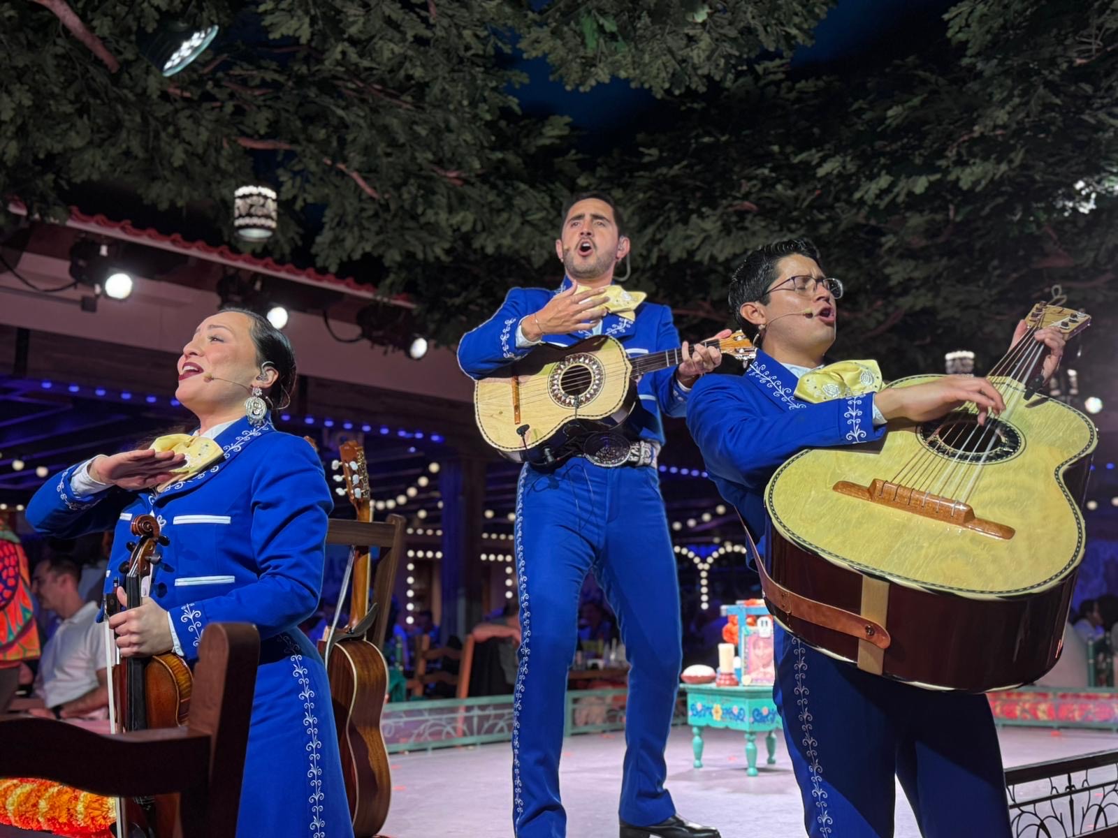 GroupPhoto Three mariachi musicians in blue outfits perform with vihuela, guitarrón, and violin on stage at Plaza de Coco aboard the Disney Treasure.