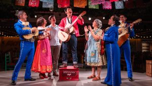 Seven people perform on a festive stage with papel picado banners, including four in mariachi outfits playing guitars.