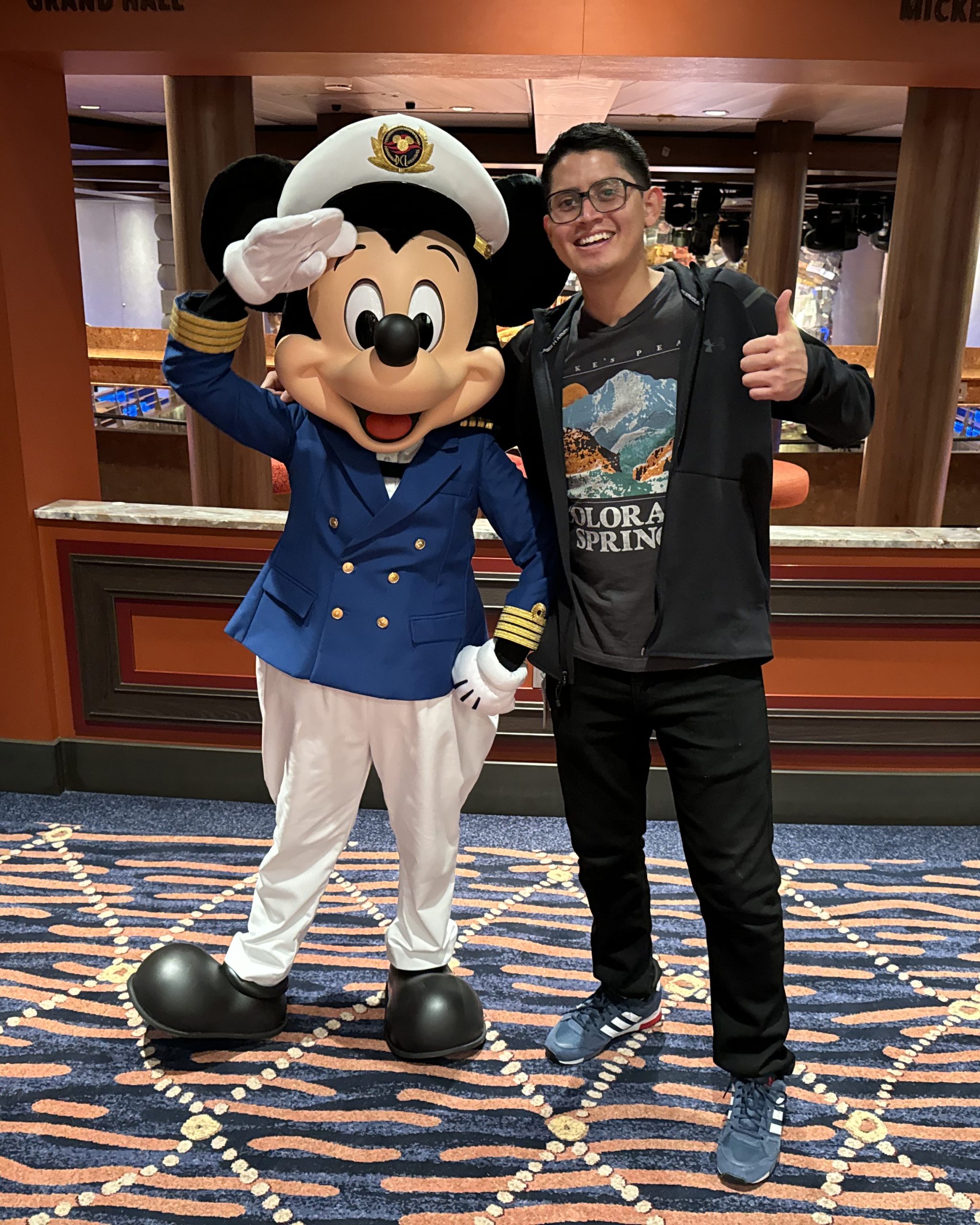 A person stands next to Mickey Mouse in a blue naval uniform, with Mickey saluting and the person giving a thumbs-up.