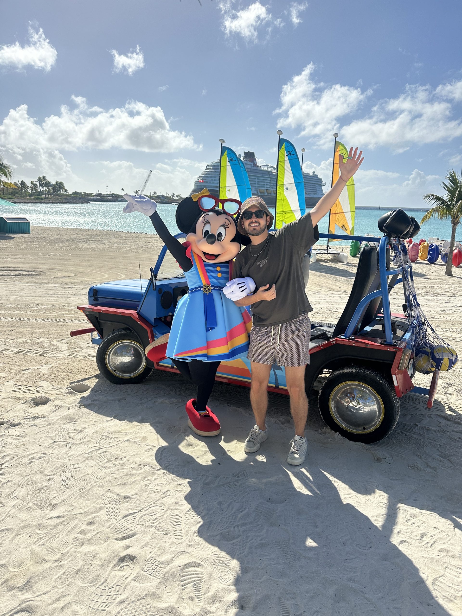A person poses with Minnie in front of a colorful dune buggy on a sandy beach with the ocean in the background.