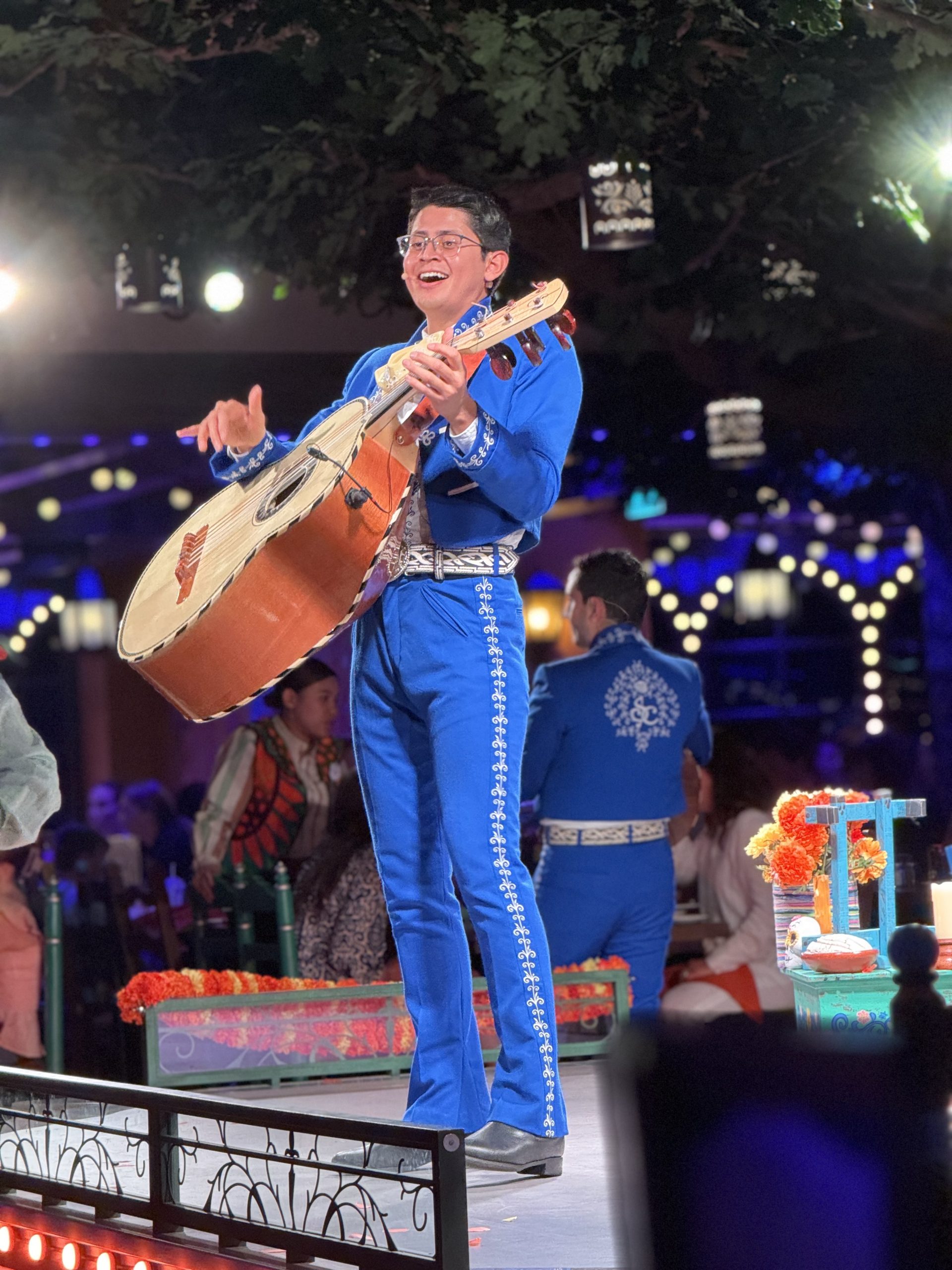 Raymundo_IMG_2911 A musician in a blue traditional mariachi outfit plays a large guitarrón on a festively decorated stage.