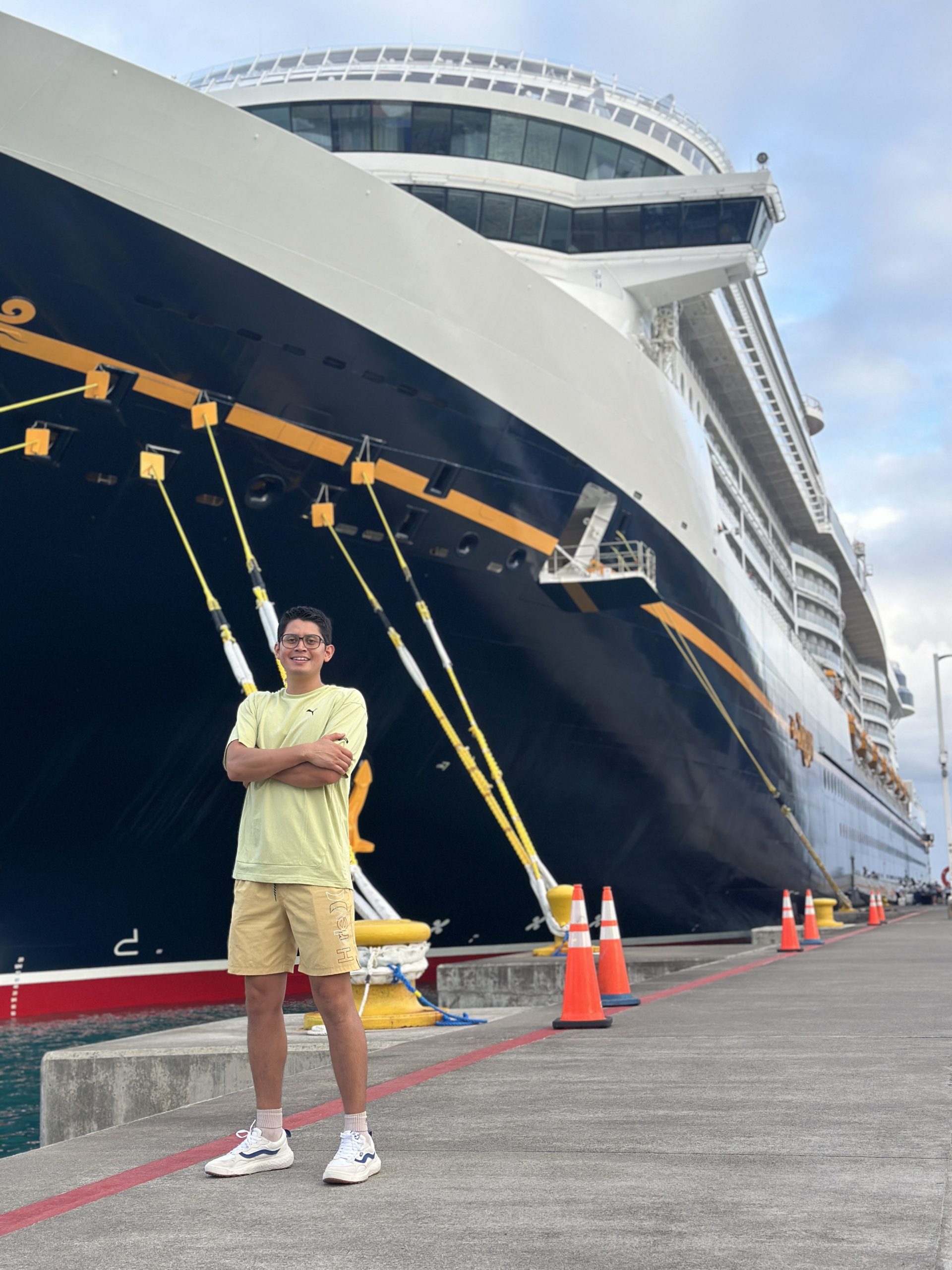 A person stands with arms crossed in front of a large cruise ship docked at a port