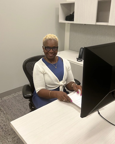 A person sits at a desk in an office, working on a computer while holding papers.