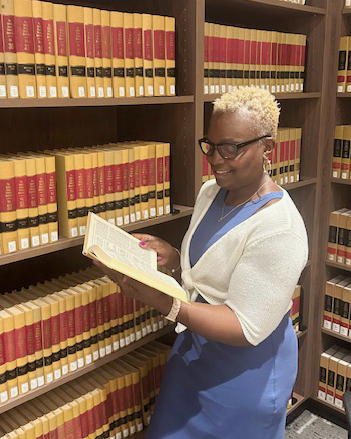 A person with short, curly hair stands in front of a bookshelf filled with yellow and red books, reading an open book.