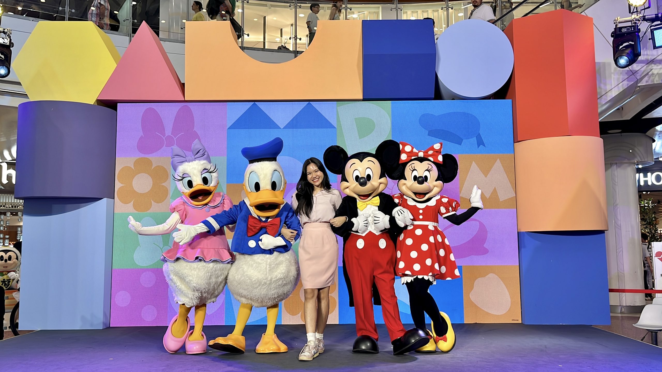 Person posing on stage with Donald Duck, Daisy Duck, Mickey Mouse, and Minnie Mouse in front of a vibrant geometric backdrop at a Disney event.