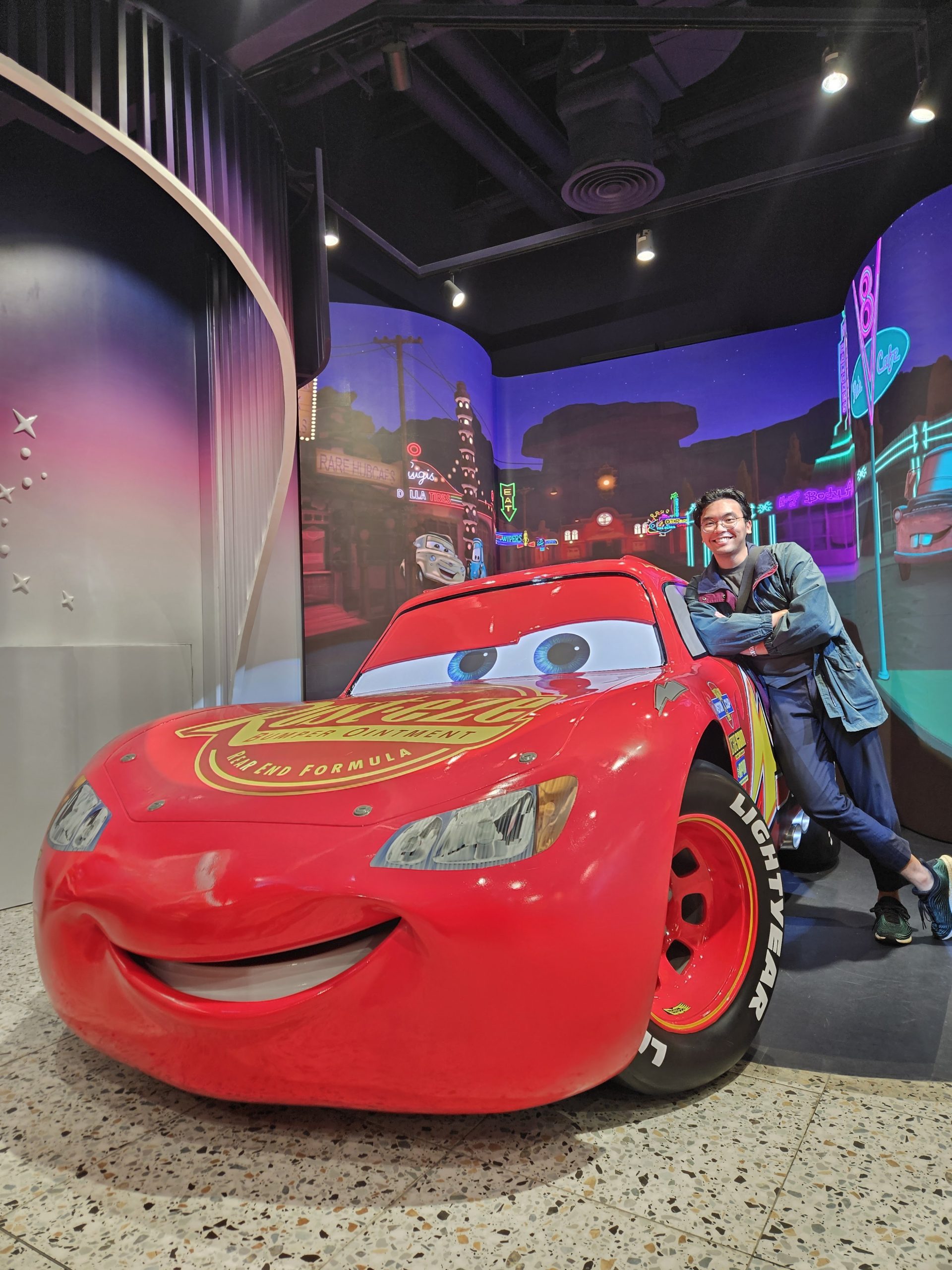 Bryan posing beside a life-sized Lightning McQueen model, set against a vibrant city-themed backdrop inspired by the movie "Cars."
