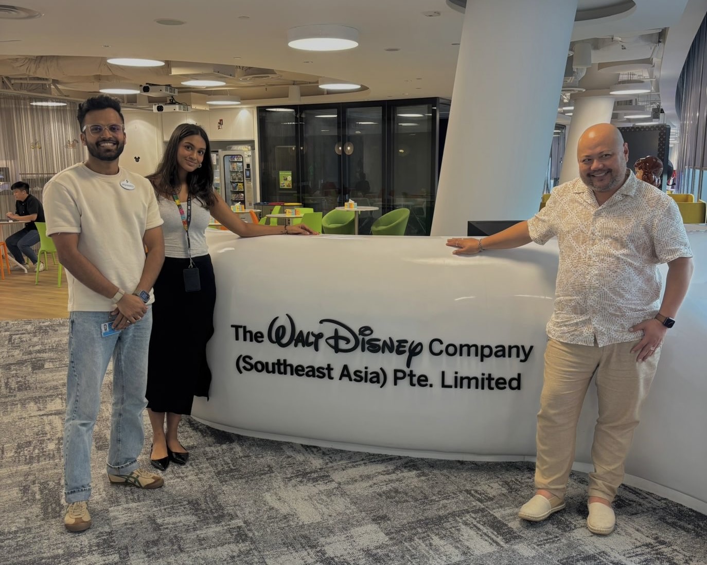 452A249D-654F-43AD-B265-3B3ACB894004_1_201_a Three people pose beside a sign for "The Walt Disney Company (Southeast Asia) Pte. Limited" in an office setting.