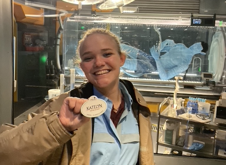 A person is holding a name tag that says "Katelyn" with show lab equipment visible in the background.
