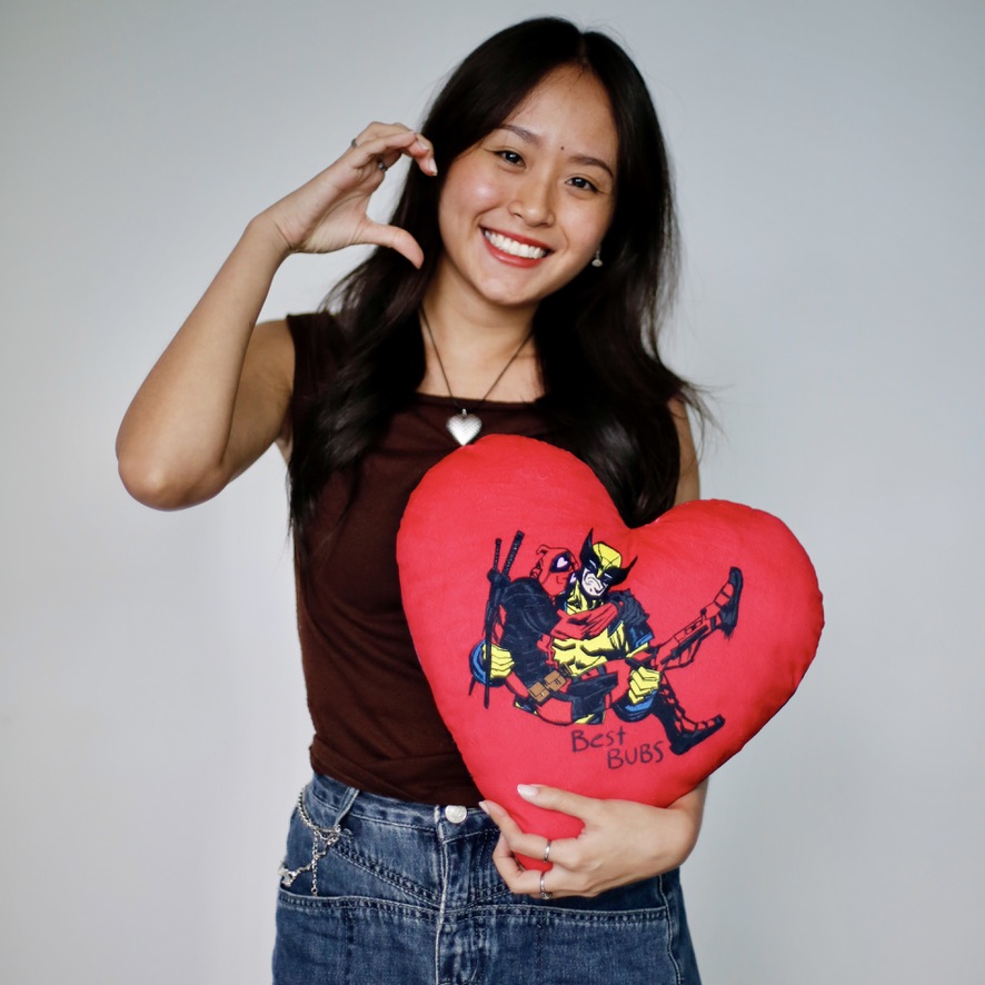 C82A57C5-0E54-4502-9F52-95E7D595F217_1_105_c A person makes a heart gesture while holding a heart-shaped pillow featuring Deadpool and Wolverine labeled “Best Bubs.”