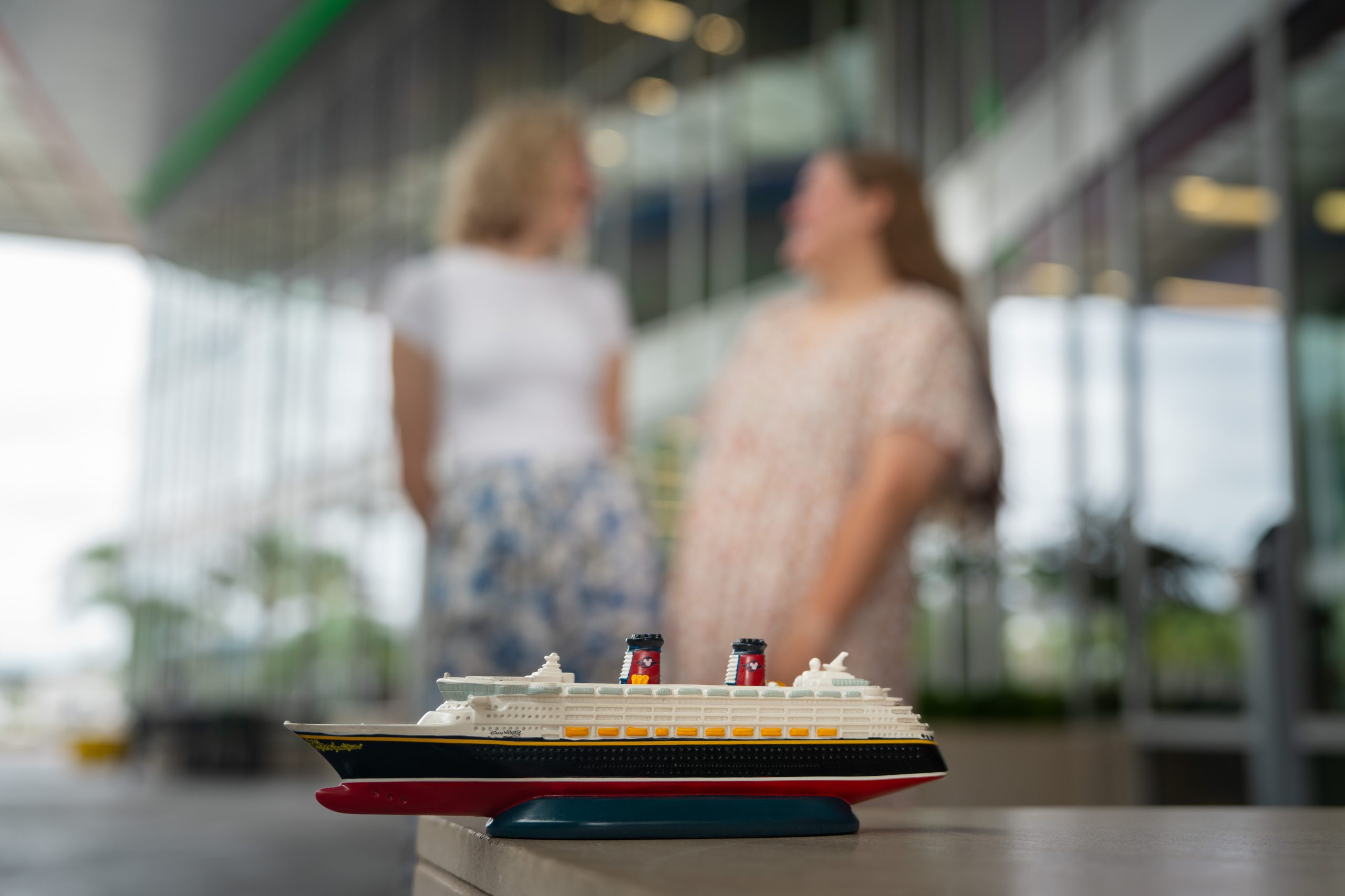 A small model of a cruise ship is displayed indoors with two blurred figures in the background.