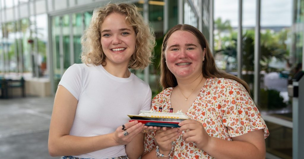 Two people are holding a small cruise ship model in front of large windows with greenery outside.