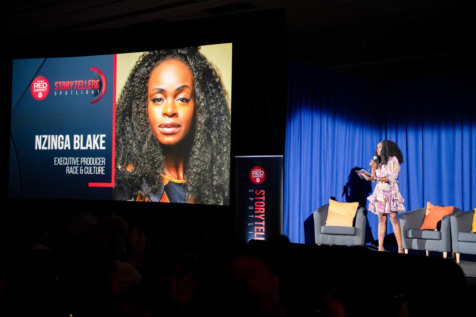 Nzinga Blake speaks on stage at the Red Storytellers Spotlight event, with a large screen displaying her name and title as Executive Producer for Race & Culture.