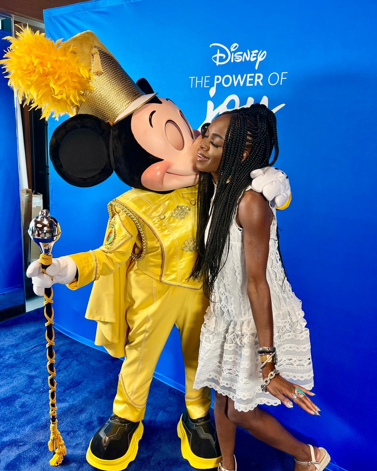 A person posing with Mickey Mouse dressed in an elaborate gold costume at a Disney event with a blue backdrop reading 'The Power of You.