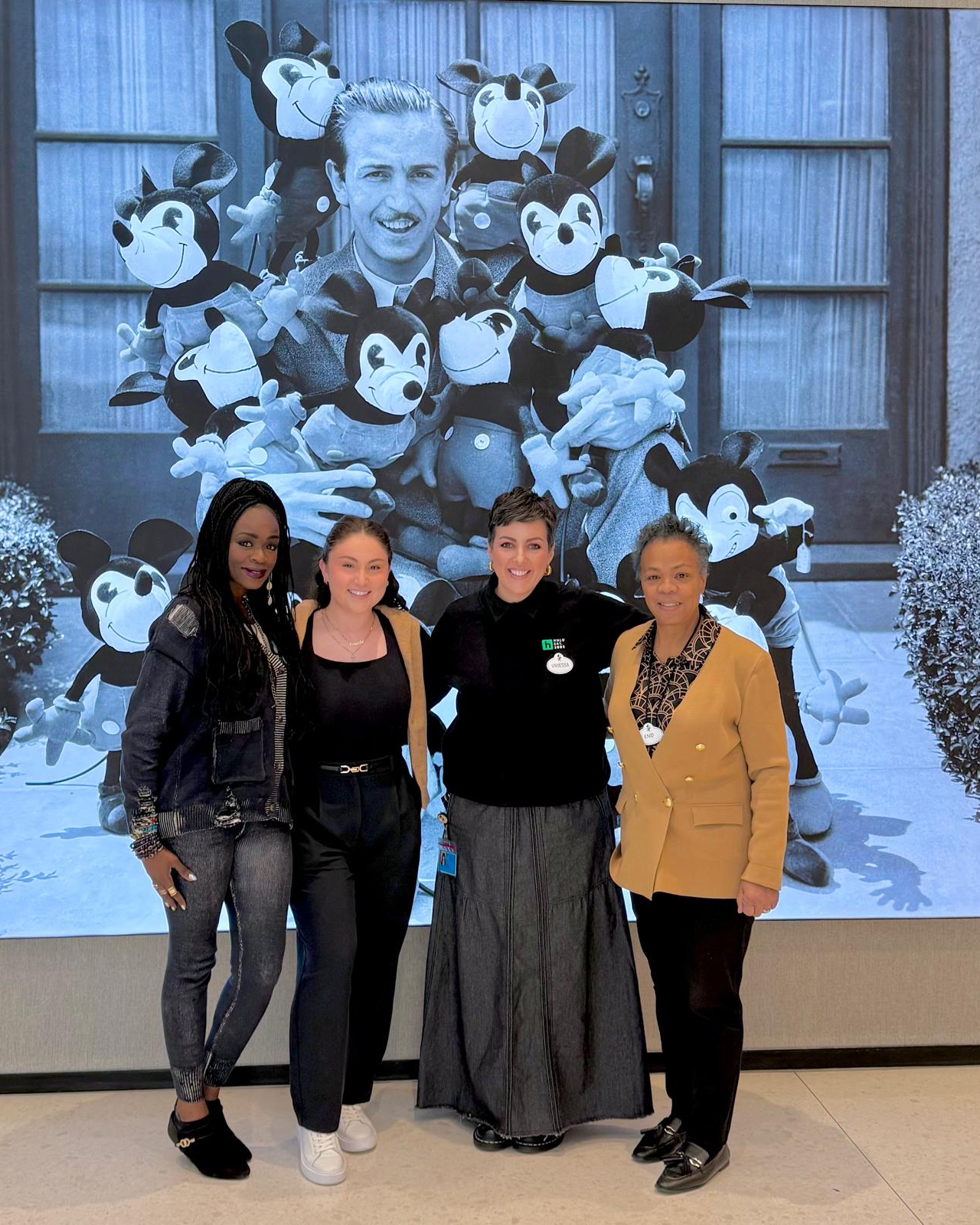 Four people standing in front of a large black-and-white photo featuring multiple classic Mickey Mouse plush toys.