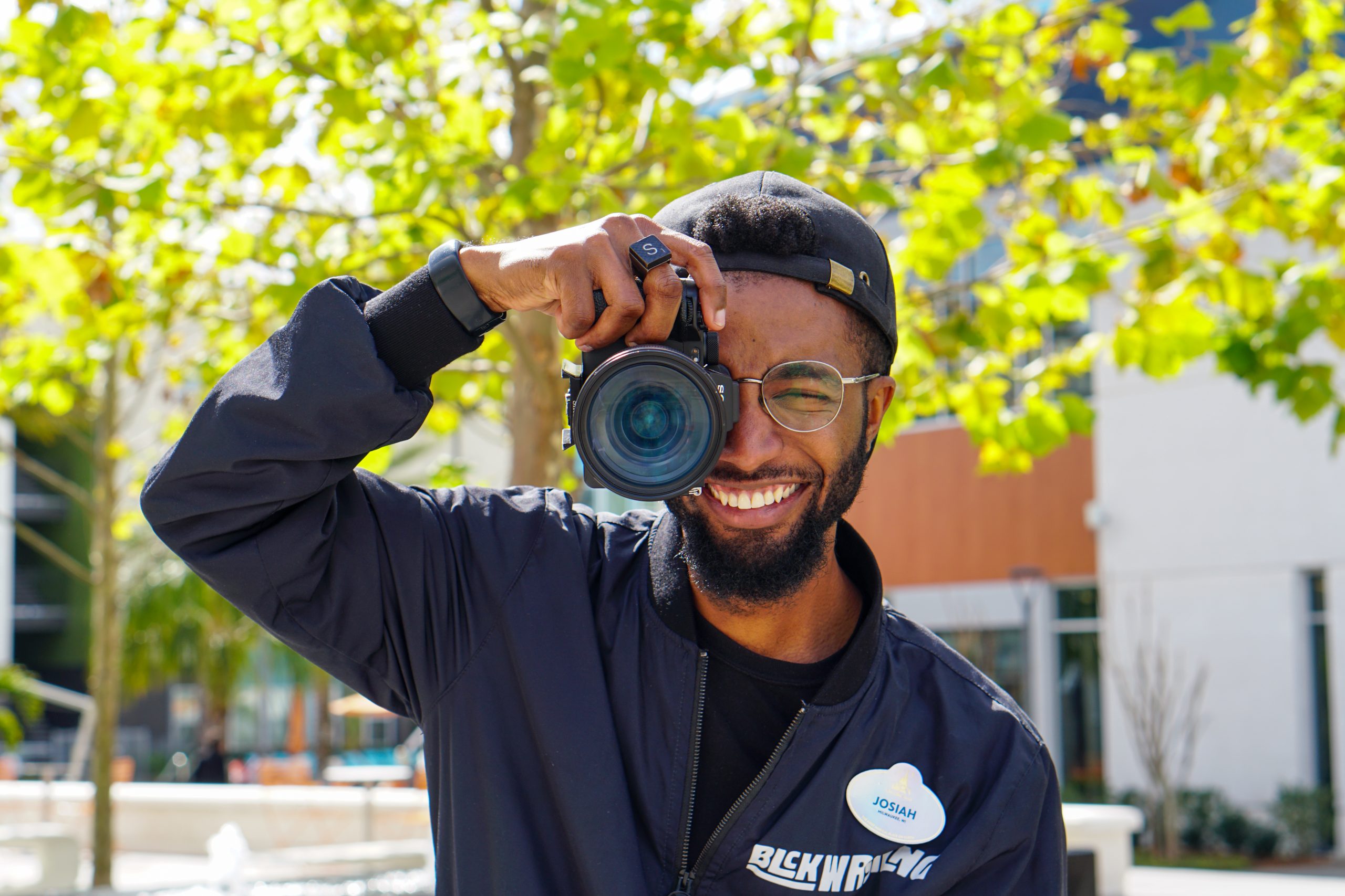 Person outdoors holding a DSLR camera up to their face, wearing a dark jacket and name badge with greenery and buildings in the background.