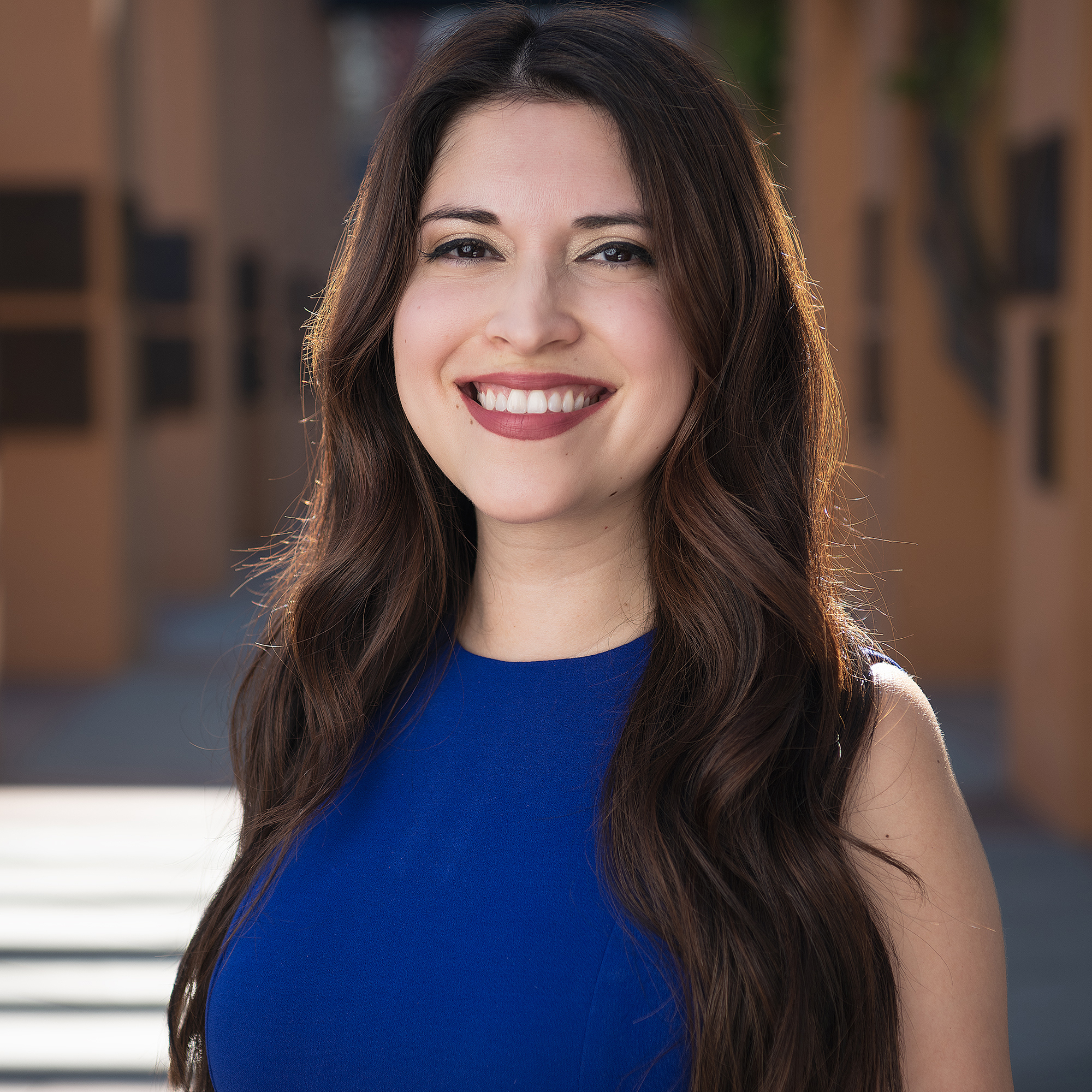 Person wearing a royal blue sleeveless dress with long wavy hair, photographed outdoors with blurred architectural background.