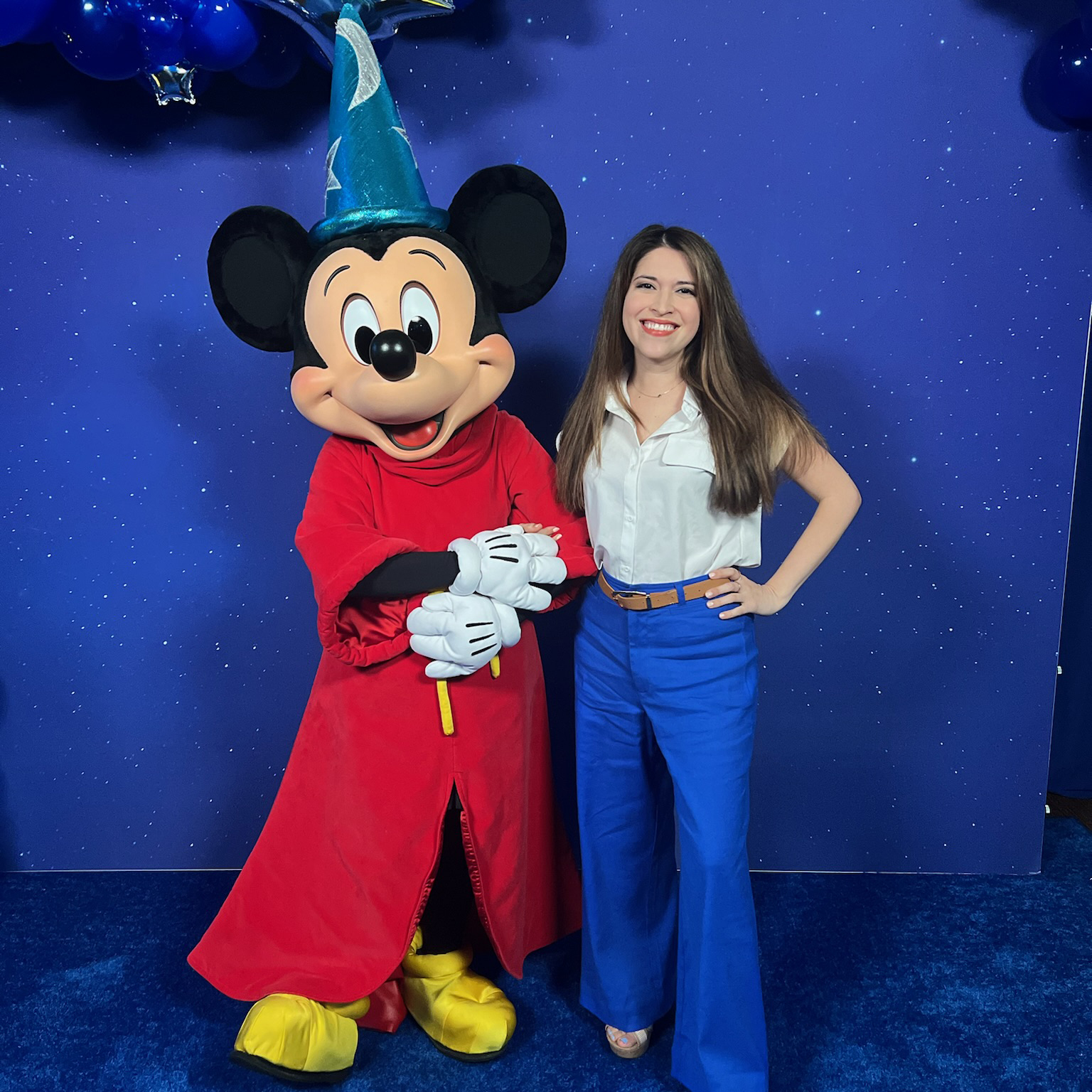 Mickey Mouse dressed as the Sorcerer’s Apprentice from Fantasia standing against a starry blue backdrop with decorative balloons.