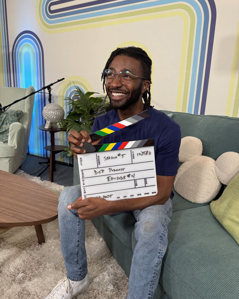 Person seated on a modern set holding a clapperboard labeled 'DCP Podcast Episode 4' with colorful retro wall design in the background.