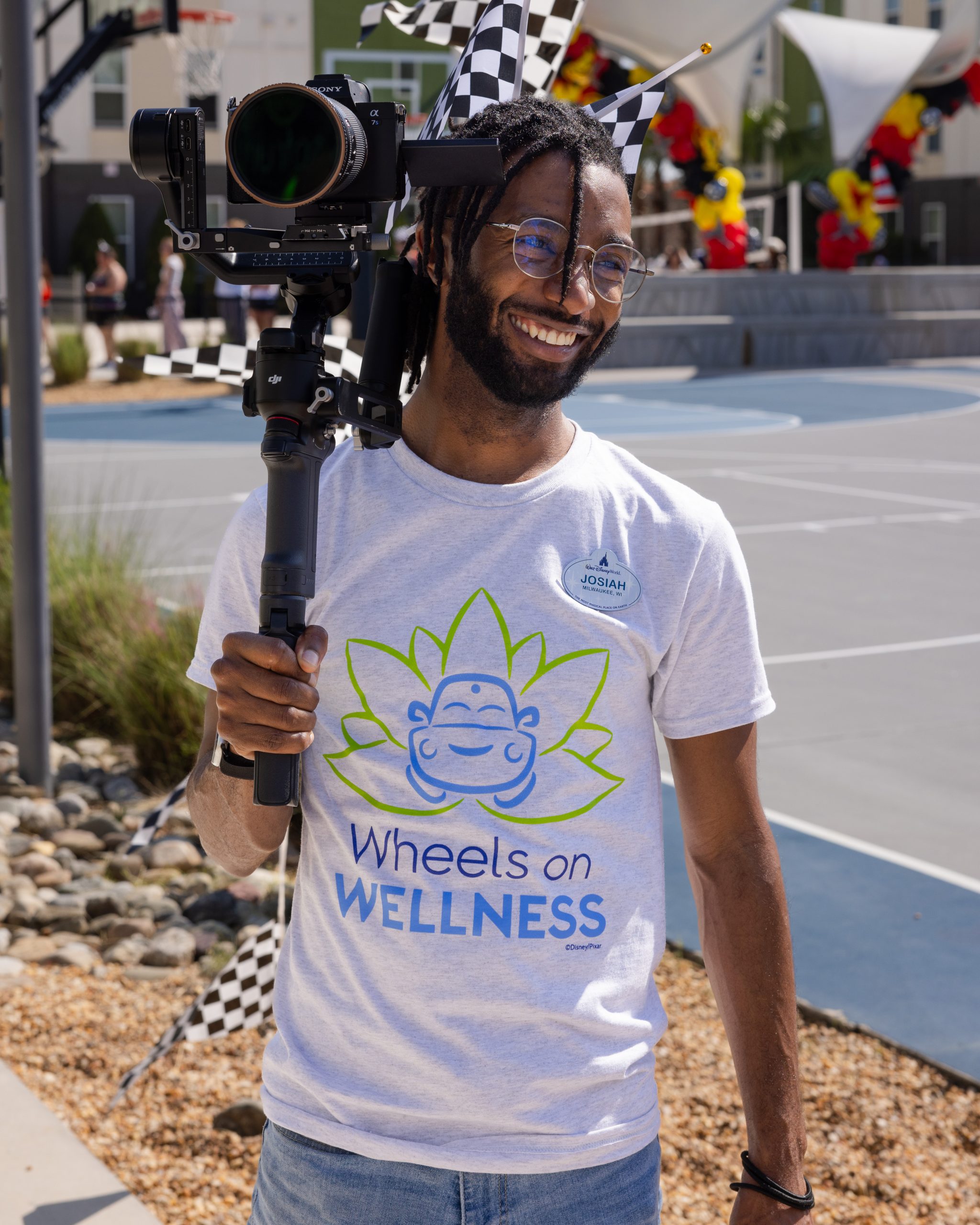Person wearing a 'Wheels on Wellness' T-shirt and holding a professional video camera at an outdoor event with checkered flags and festive decorations.