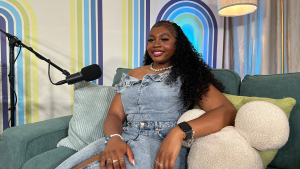 Person in denim sits on green couch with microphone, colorful striped background, and decorative pillows.