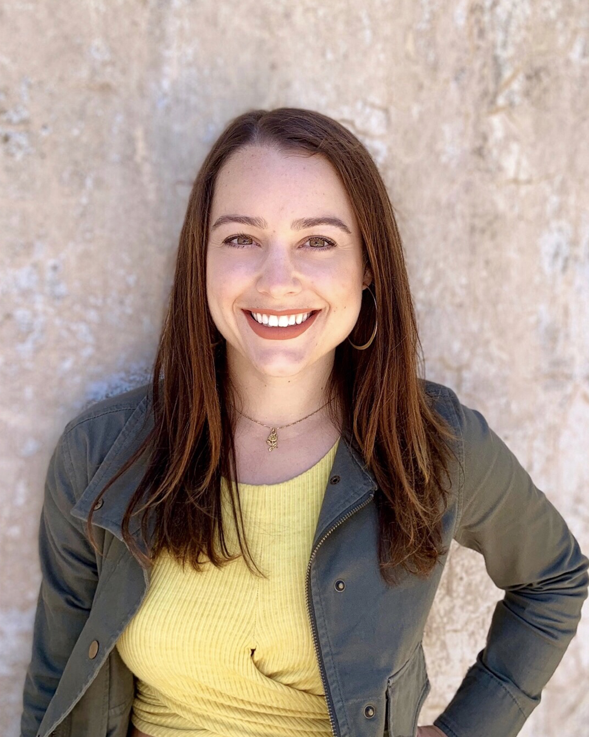 Person wearing a yellow top and green jacket standing against a textured beige wall in natural light.