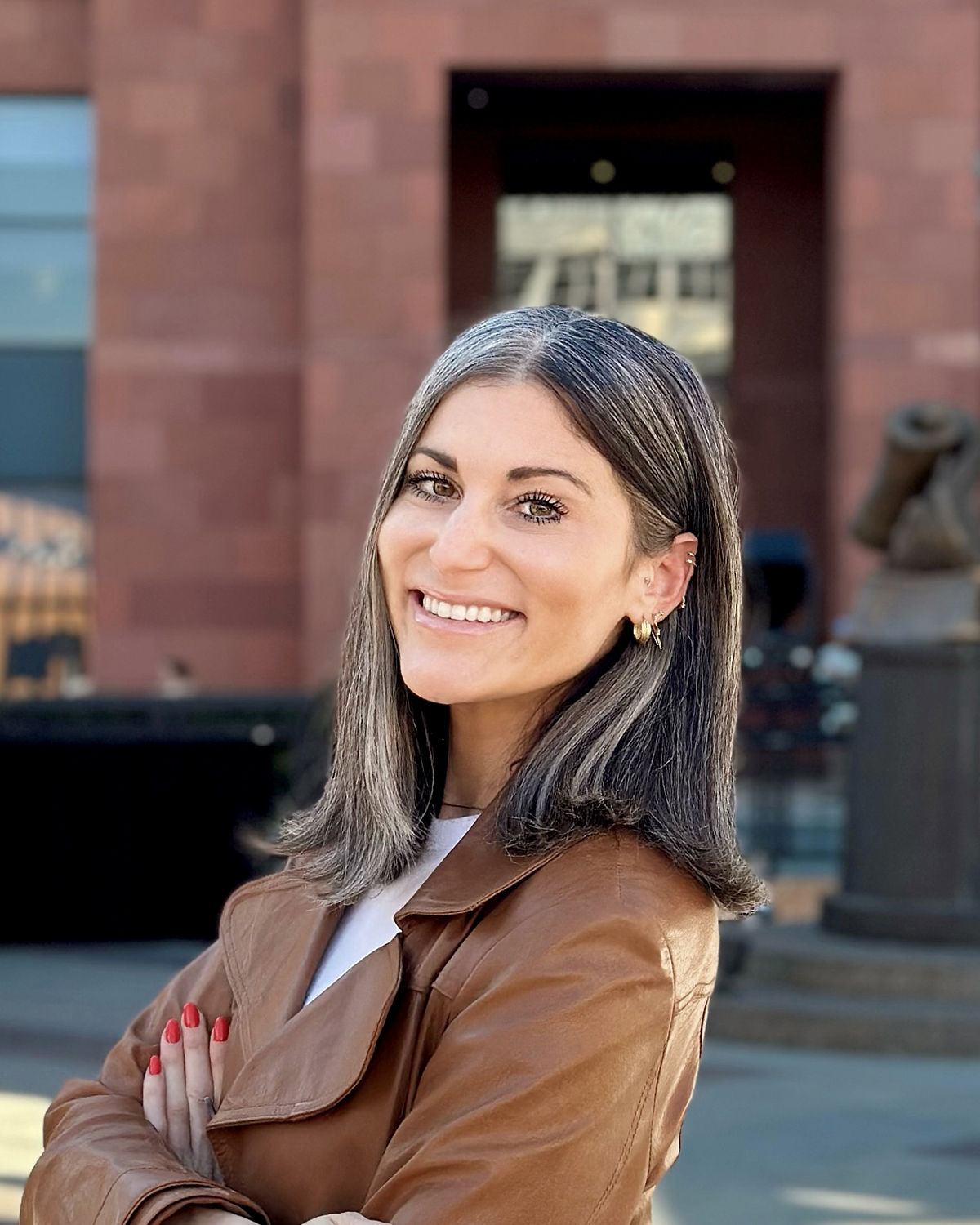 Person wearing a brown leather jacket with arms crossed, standing outdoors in front of a red brick building and a blurred statue in the background.