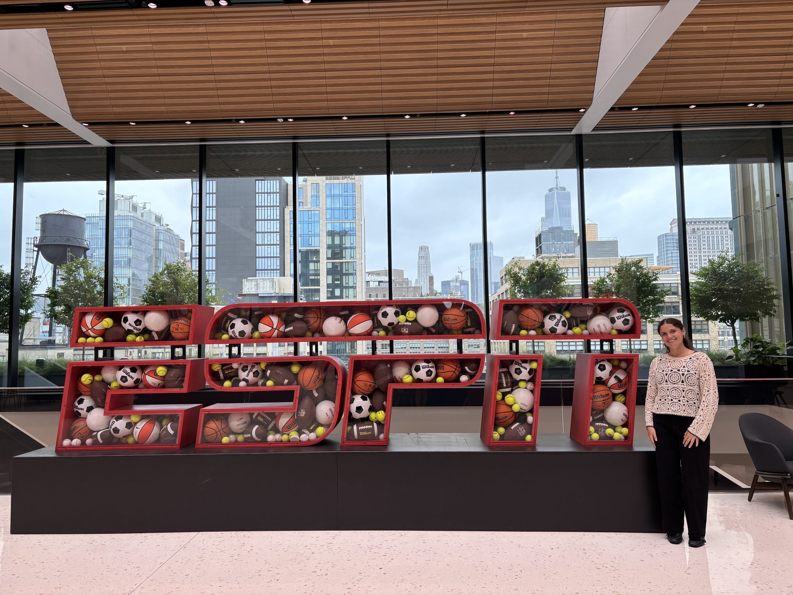 Person standing next to a large ESPN sculpture filled with sports balls in a modern office space, symbolizing ESPN’s diverse sports coverage and creative workplace.