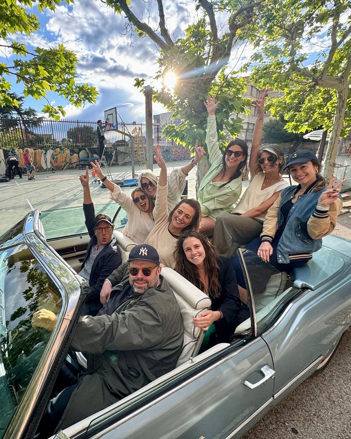 Group of people sitting and standing in a vintage convertible car outdoors, raising their arms in celebration under a sunny sky with trees and a basketball court in the background.