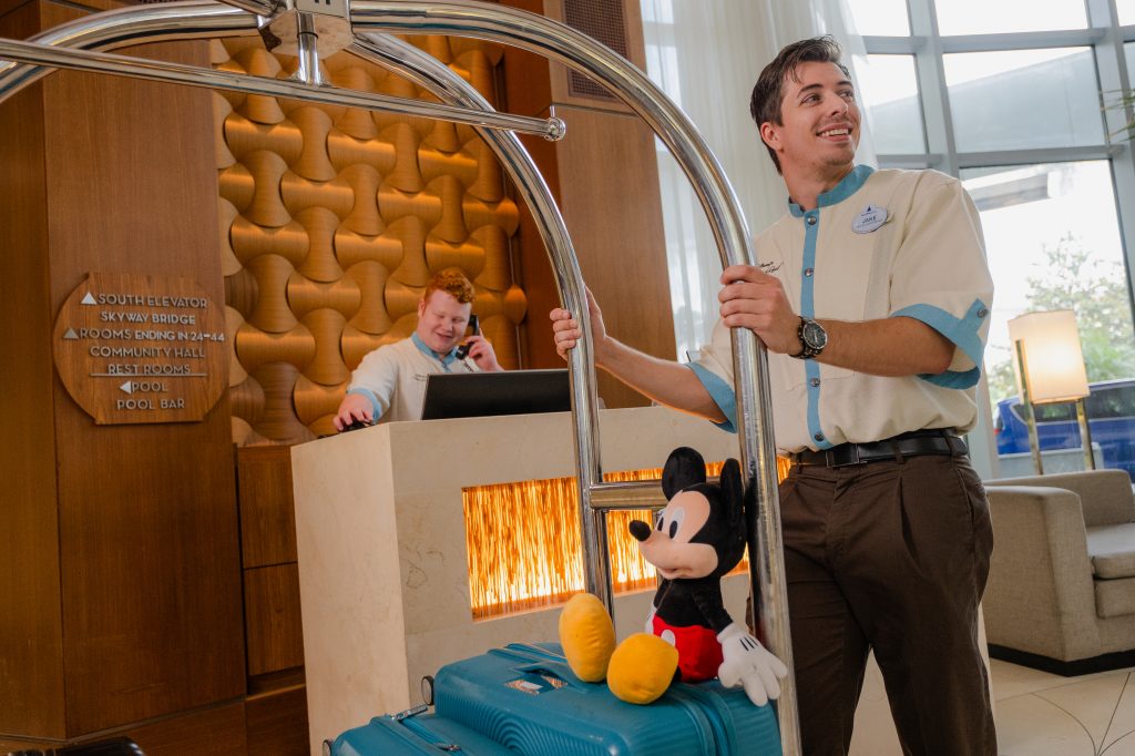 Bell services staff holding a luggage cart with a suitcase and a Mickey Mouse plush in a hotel lobby near a reception desk.