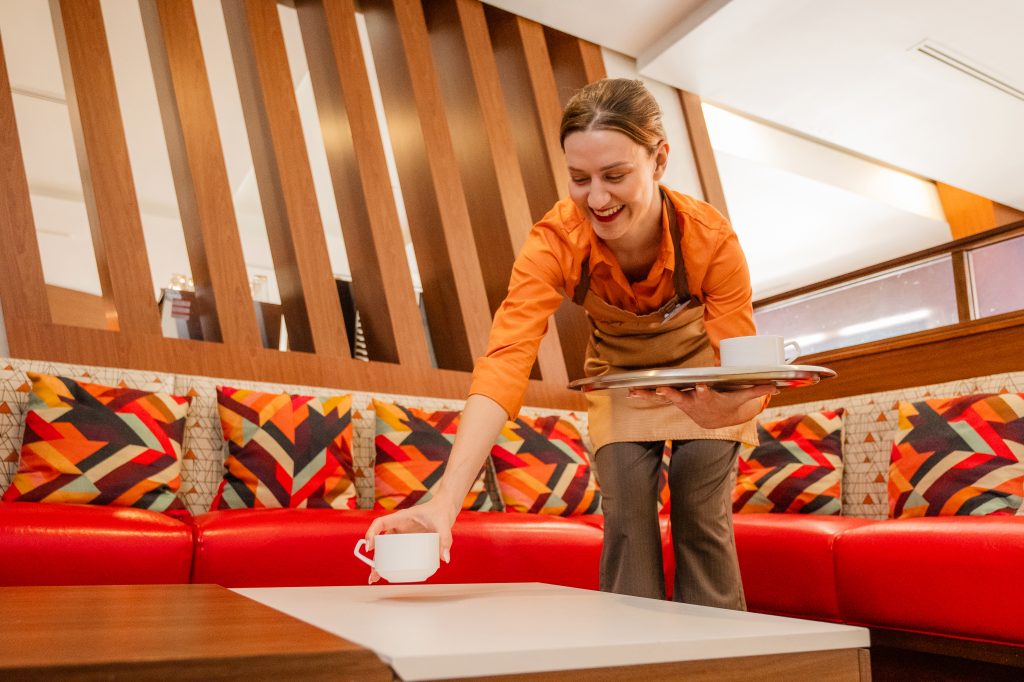 A person in an apron placing a cup on a table while holding a tray in a lounge with colorful patterned cushions.