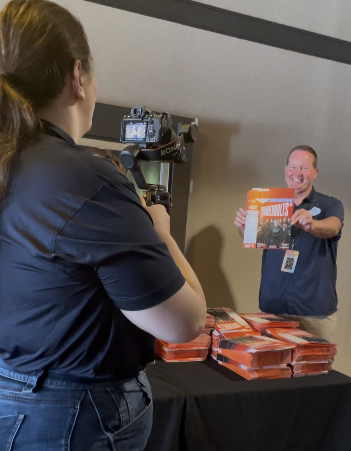 Person filming another person holding an orange booklet titled &ldquo;The Thunderbolts&rdquo; in front of a table stacked with similar booklets.