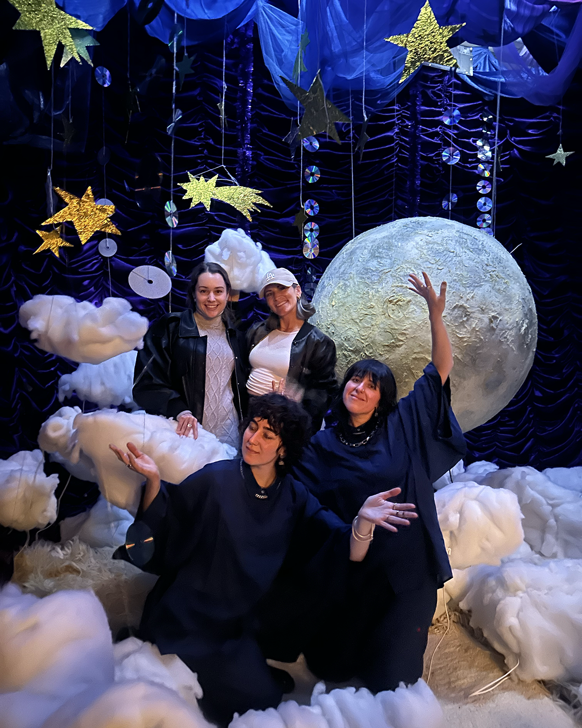 Four people posing in a whimsical holiday display featuring fluffy clouds, a large moon prop, hanging stars, and shimmering decorations against a deep blue backdrop.