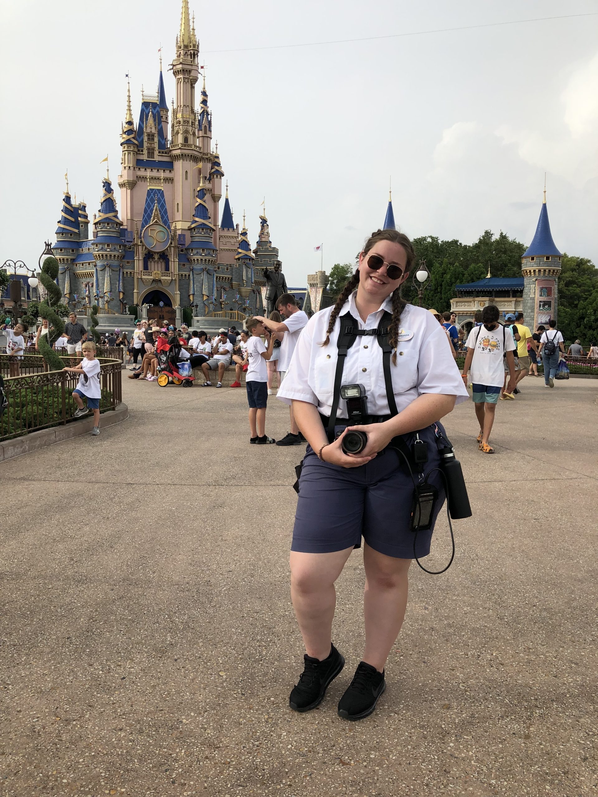 Person in a white uniform holding a camera in front of a castle during the day with crowds in the background.