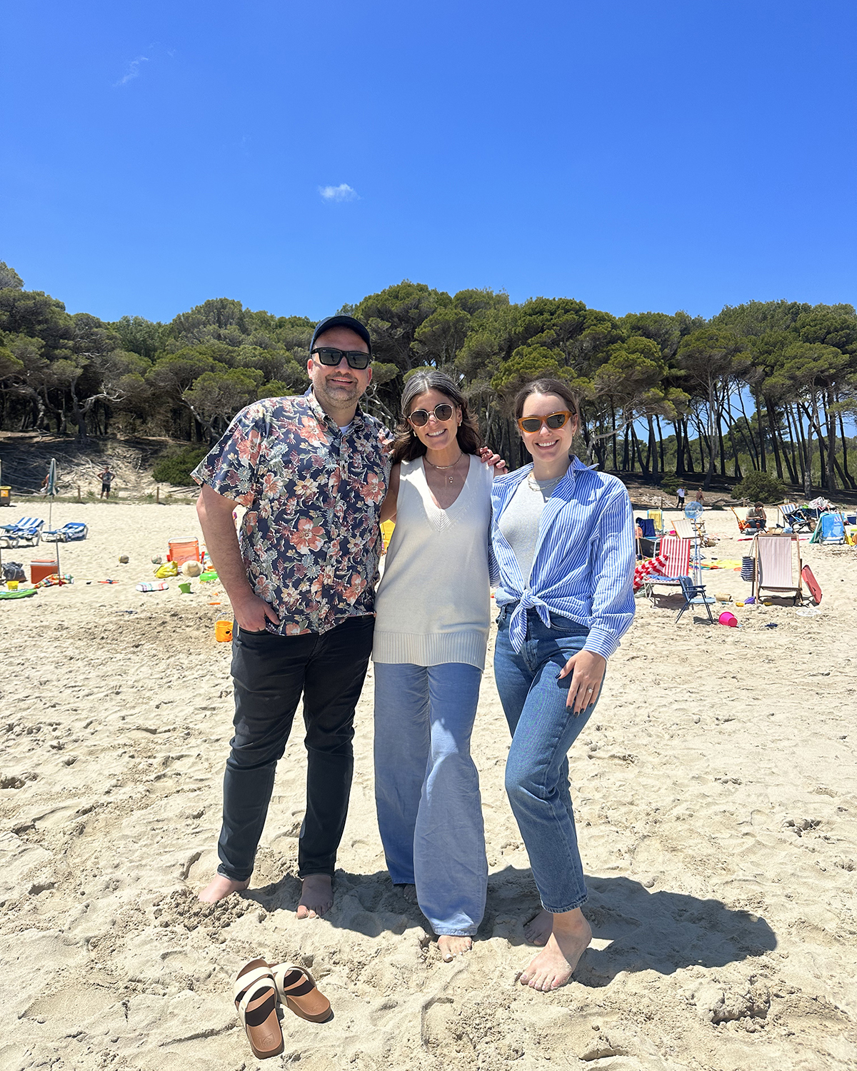 Three people standing barefoot on a sandy beach with colorful towels and toys scattered around, lush green trees in the background, and a bright blue sky overhead.