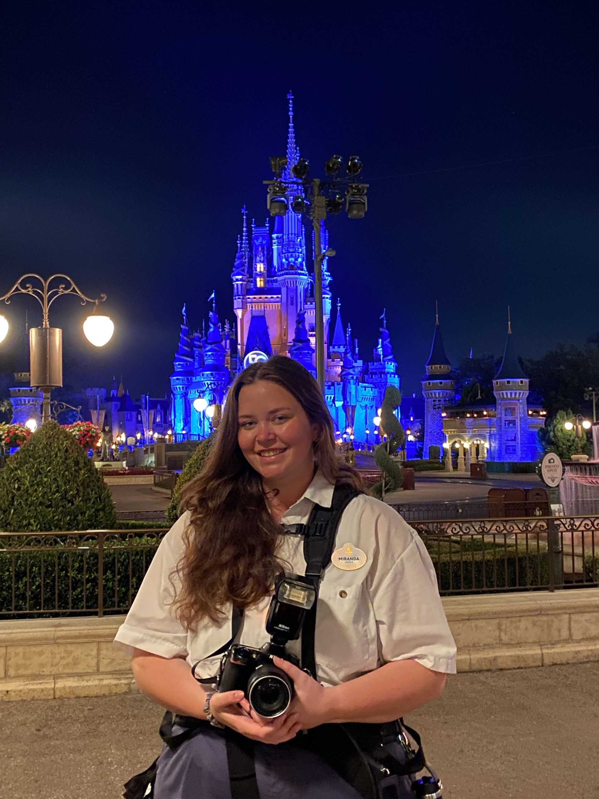 Person in a white uniform holding a camera in front of an illuminated castle at night.