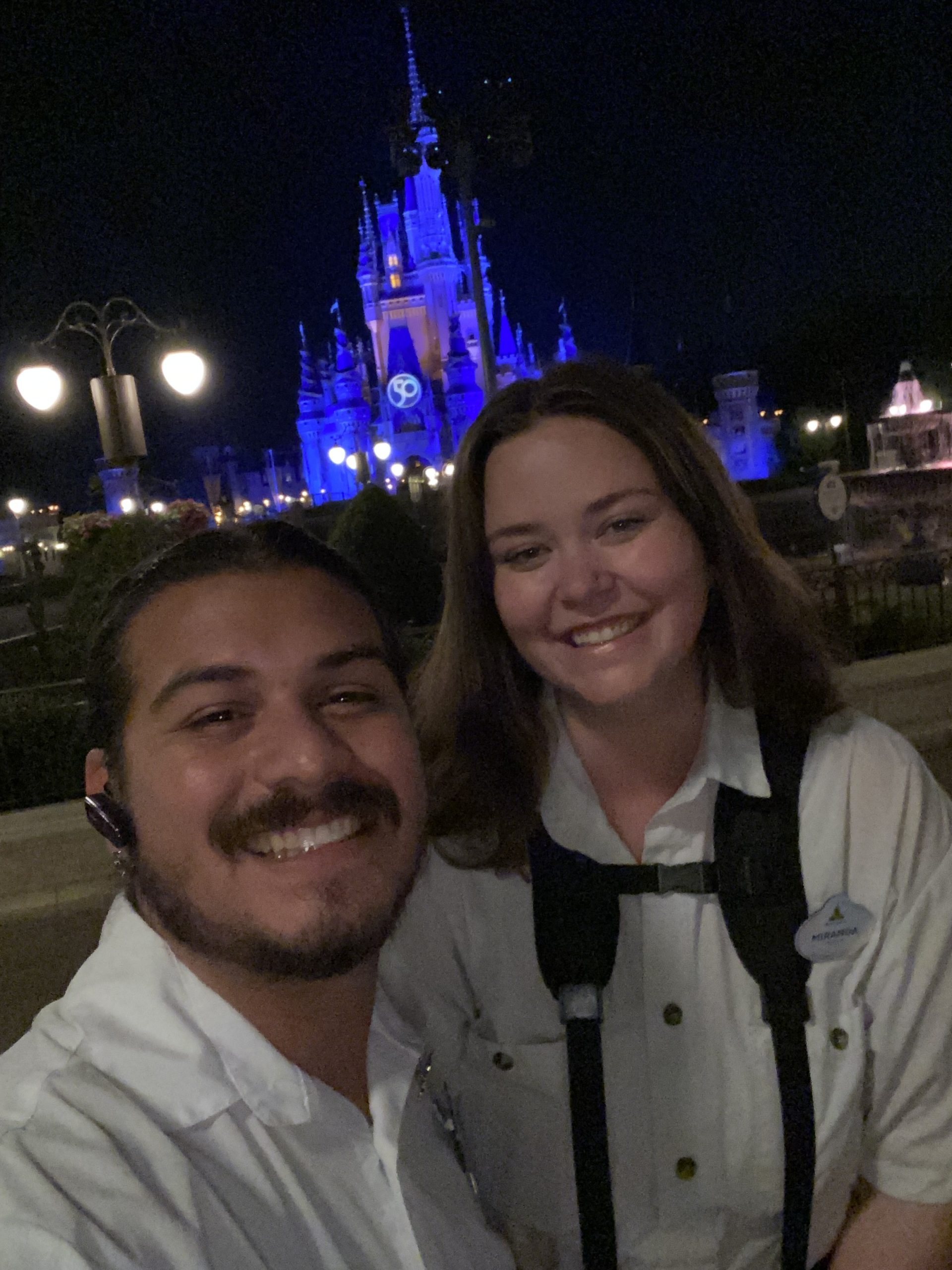 Two people in white uniforms posing in front of an illuminated castle at night.