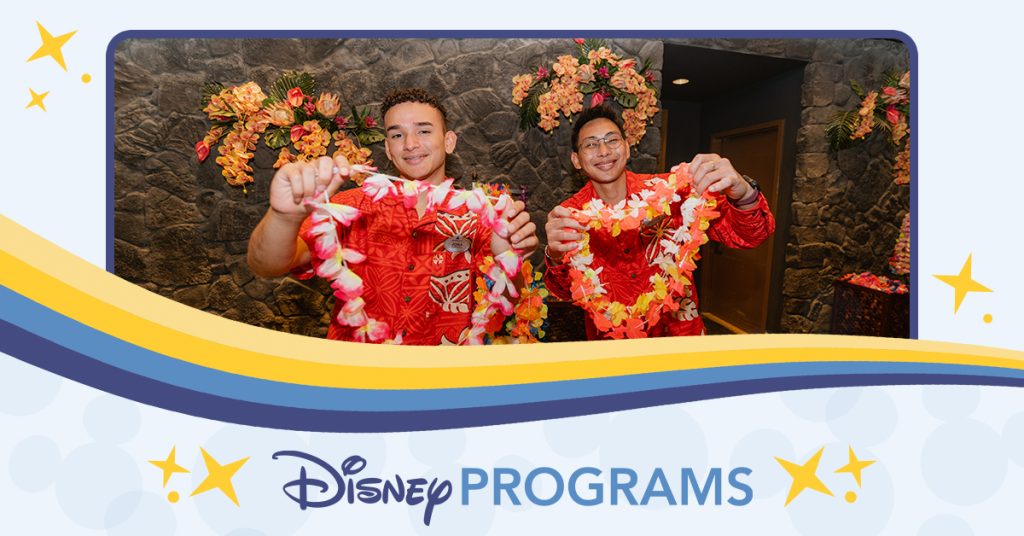 Two people in red floral shirts holding colorful leis behind a counter decorated with tropical flowers.