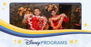 Two people in red floral shirts holding colorful leis behind a counter decorated with tropical flowers.