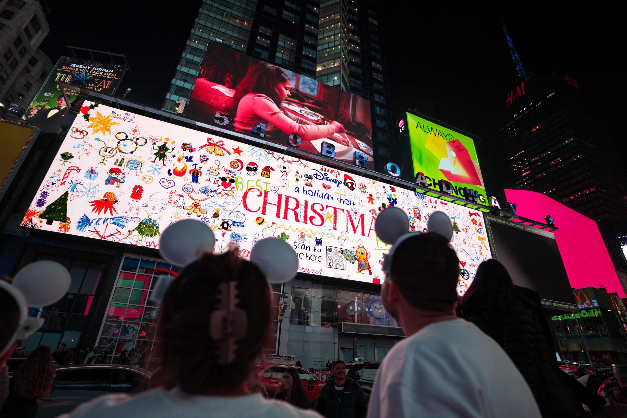 People wearing Mickey Mouse ears looking up at a bright Times Square billboard promoting Disney&rsquo;s holiday short Best Christmas Ever, surrounded by colorful lights and skyscrapers.
